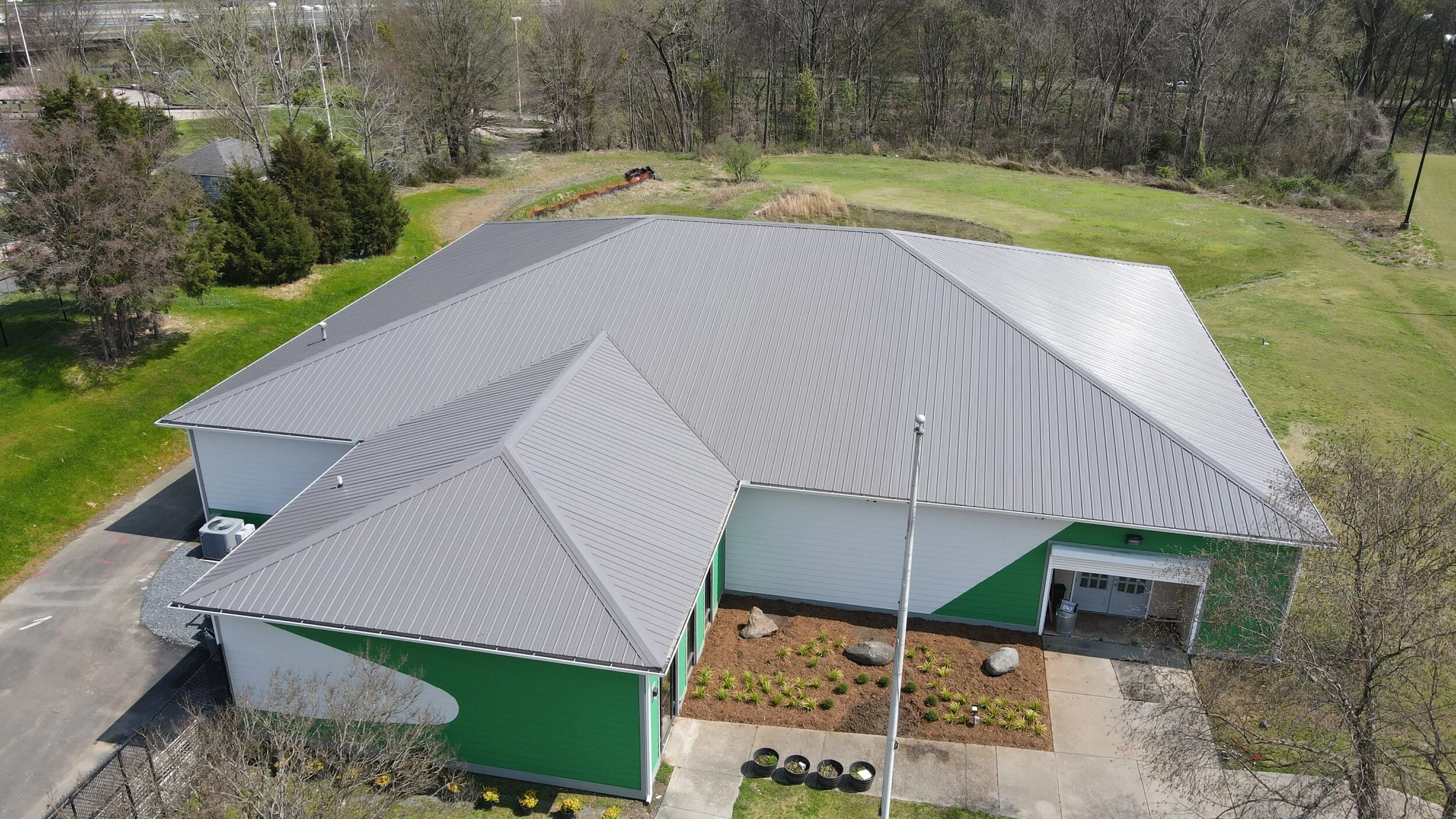 Aerial view of a modern building with green and white exterior walls, metal roof, and surrounding landscaped area with small plants, rocks, and trees.