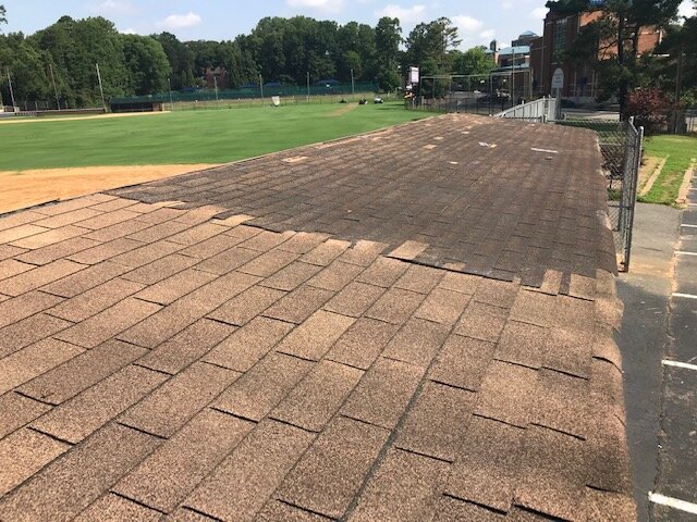 A rooftop with asphalt shingles, some of which are damaged or missing, overlooking a sports field with green grass and trees.