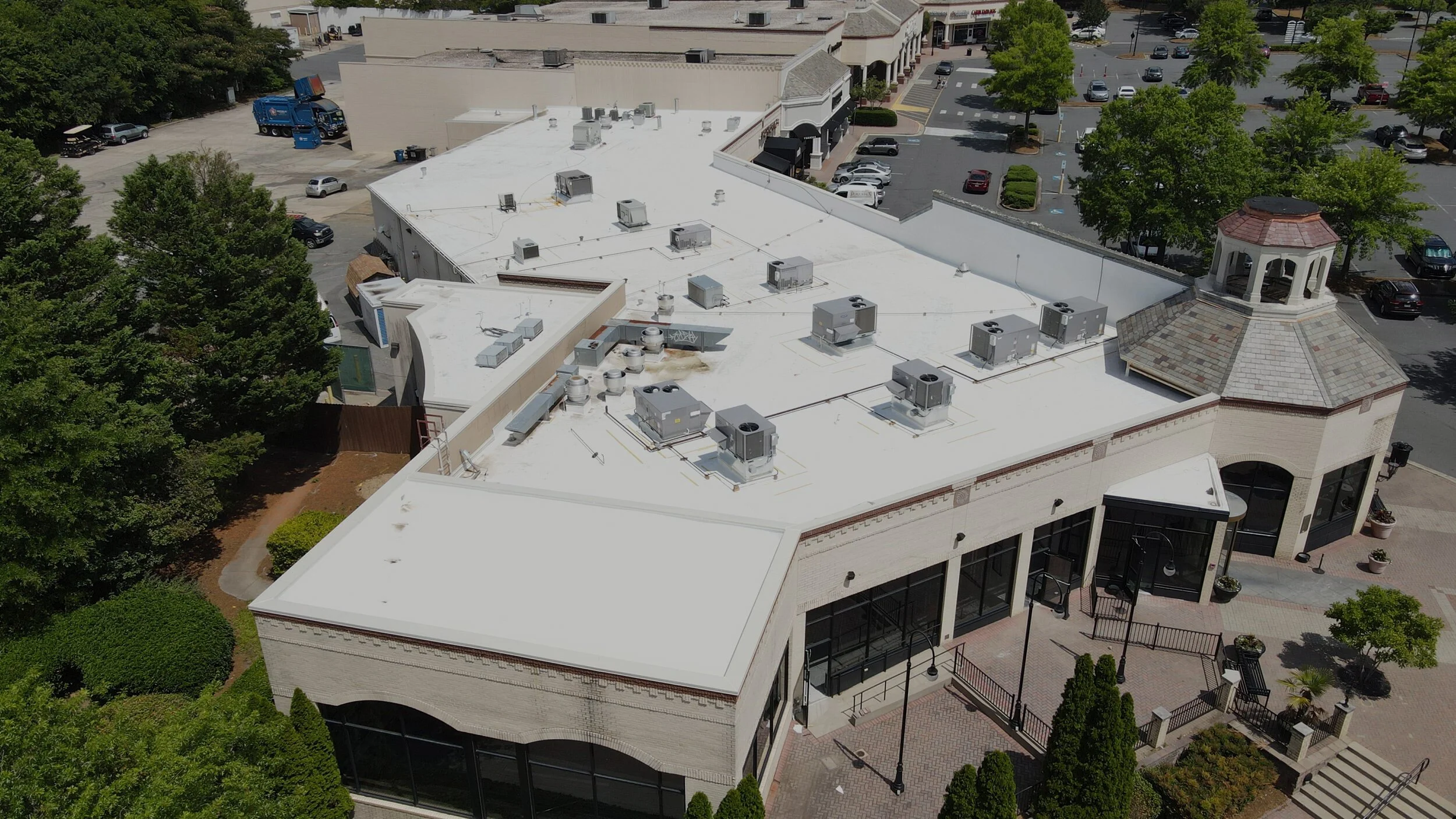 Aerial view of a shopping mall with a white flat roof and a small tower. Surrounding the mall are parking lots with cars and green trees.