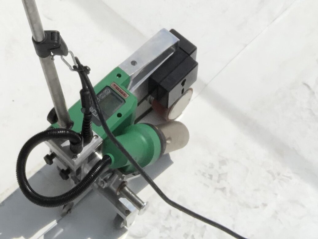 Close-up view of a piece of industrial equipment or machinery with various components, including a green box, metallic parts, black and gray elements, and a black power cord, situated on a white surface.