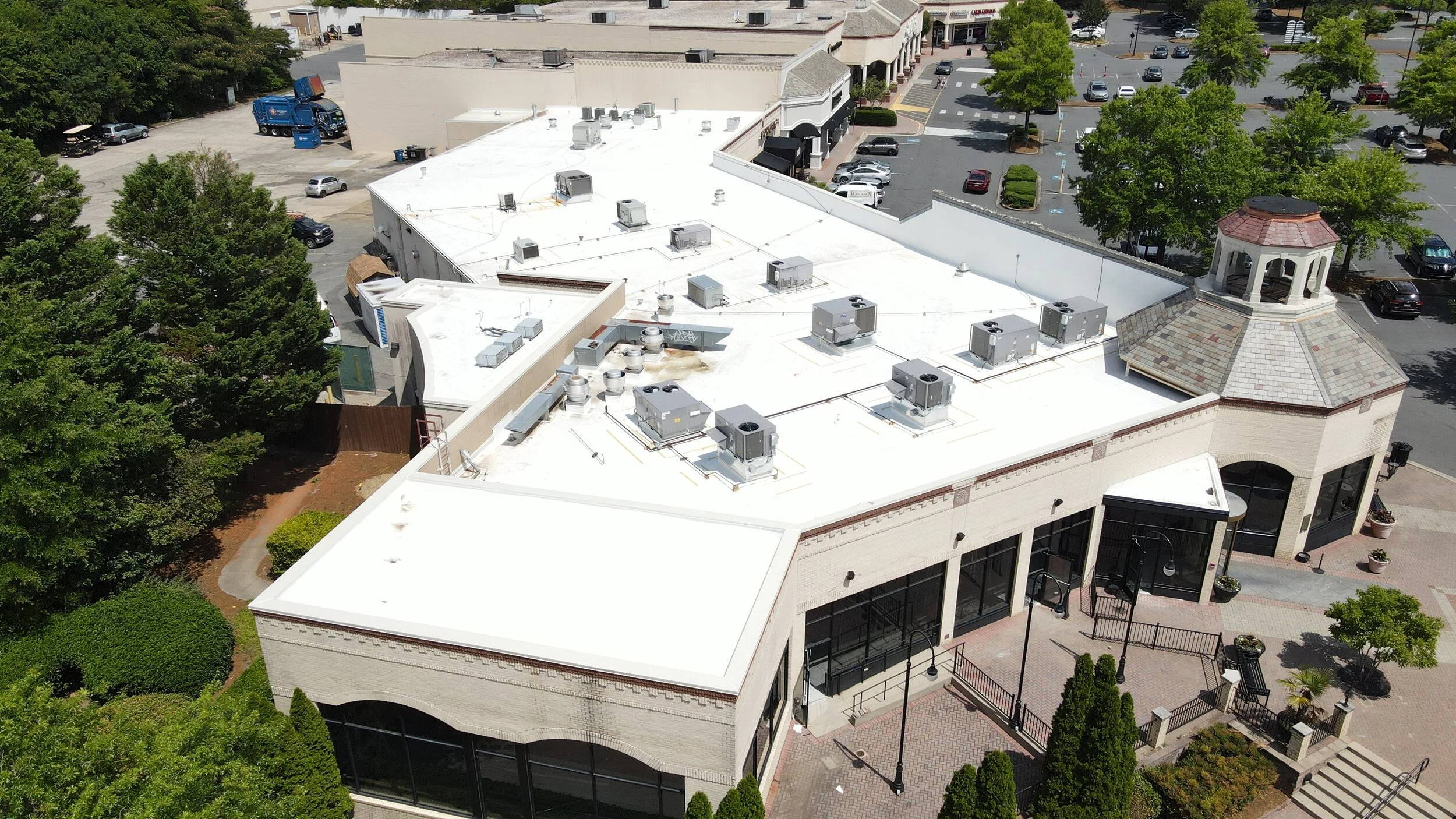 Aerial view of a shopping mall with a white roof, trees, parking lot, and a small octagonal tower with a red roof.