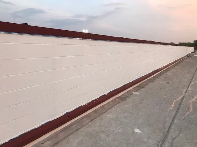 Long beige brick wall with red trim near the roof and bottom, extending along a concrete sidewalk with some cracks, under a cloudy sky.