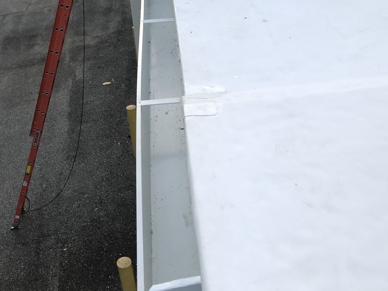 Close-up of a construction site showing a white flat surface, possibly a roof or wall, with a red ladder leaning against a dark paved ground and wooden posts supporting the structure.