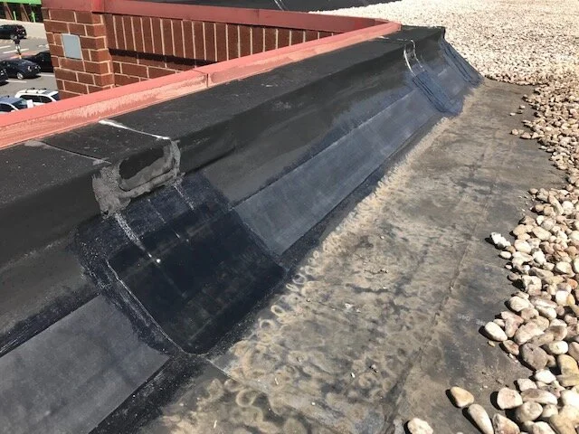 Close-up of a rooftop with a black rubber membrane, sealing seams and flashing, surrounded by gravel on one side and brick building in the background.
