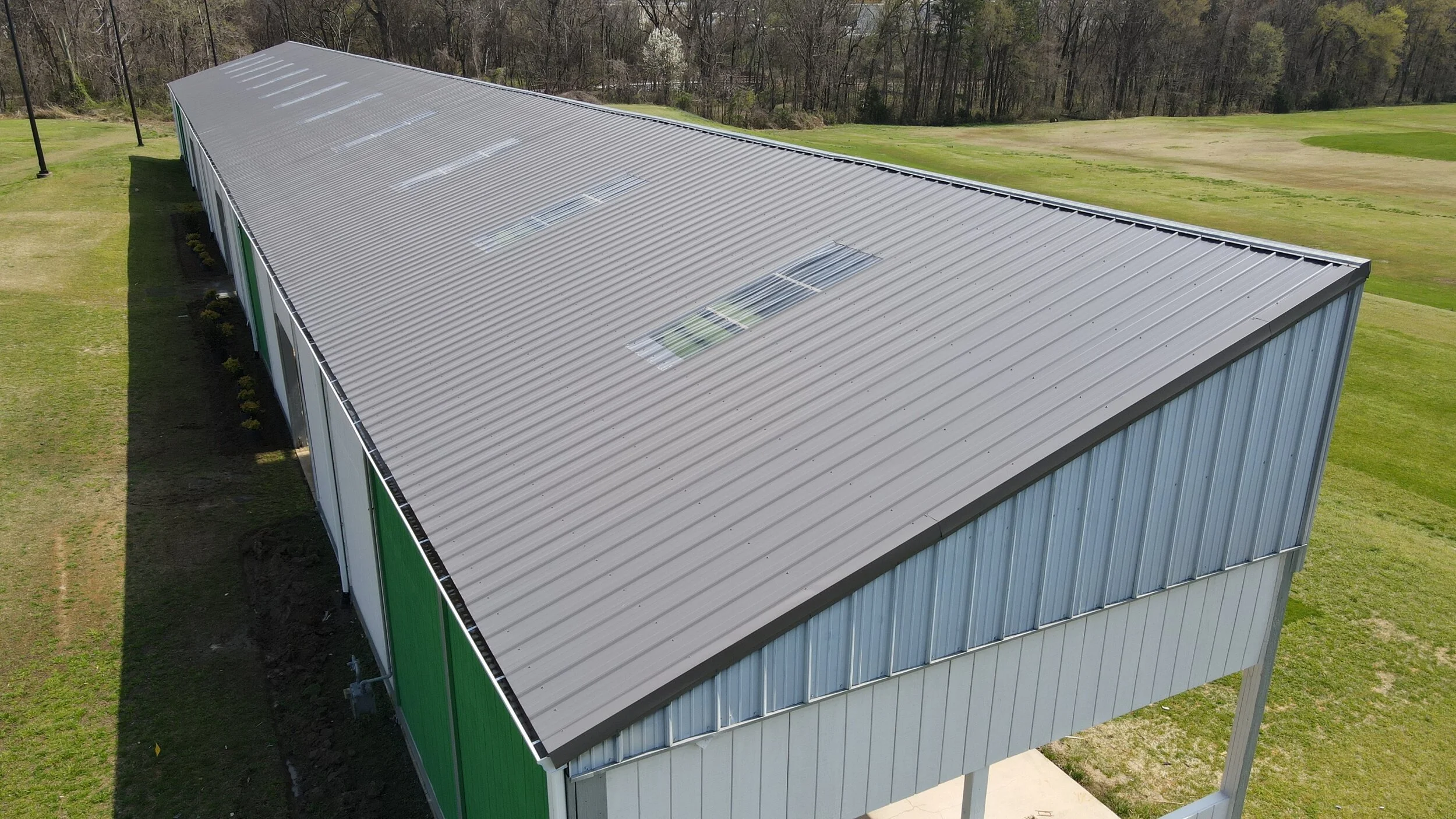 An aerial view of a large, rectangular metal building with a silver roof and green and gray walls, situated on a grassy field with some trees in the background.