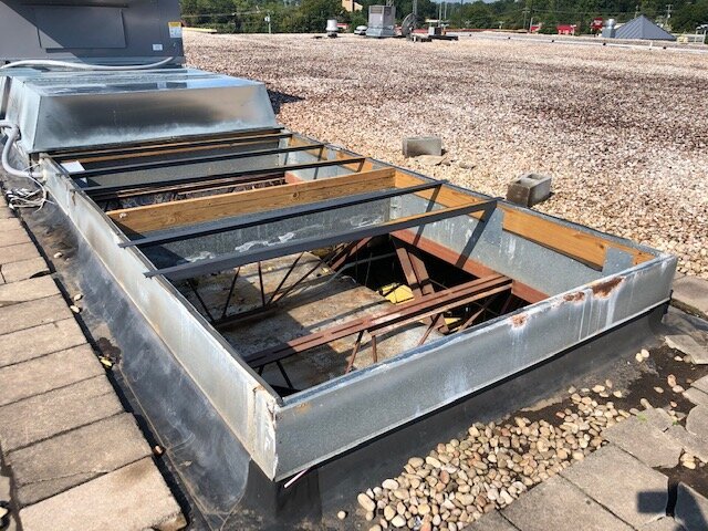 A rooftop skylight with a metal frame and glass panels, some panels have fallen out, exposing the interior supports. The skylight is on a flat roof with gravel and some concrete blocks nearby.
