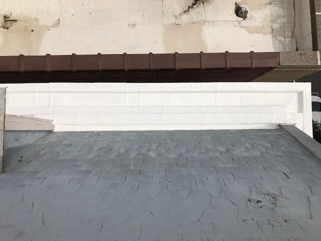 View of a building's rooftop showing a gray flat roof surface with cracked paint in the foreground and a white wall with horizontal lines in the background, along with a brown gutter along the roof's edge.