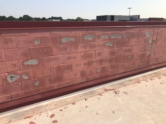 A red brick wall with several patches of peeling paint or damage and a metal or plastic railing on top. The wall is on a rooftop or outdoor area with a concrete surface and some scattered debris, with trees and buildings in the background.