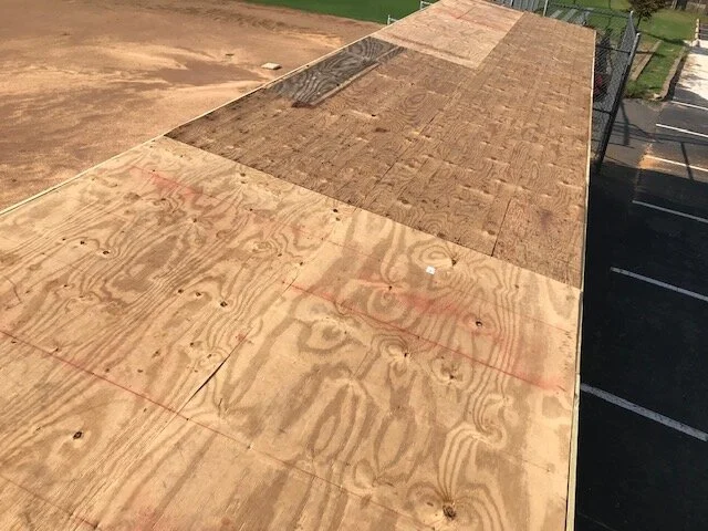 Wooden plywood sheets covering a flat rooftop, with a baseball diamond and parking lot visible in the background.
