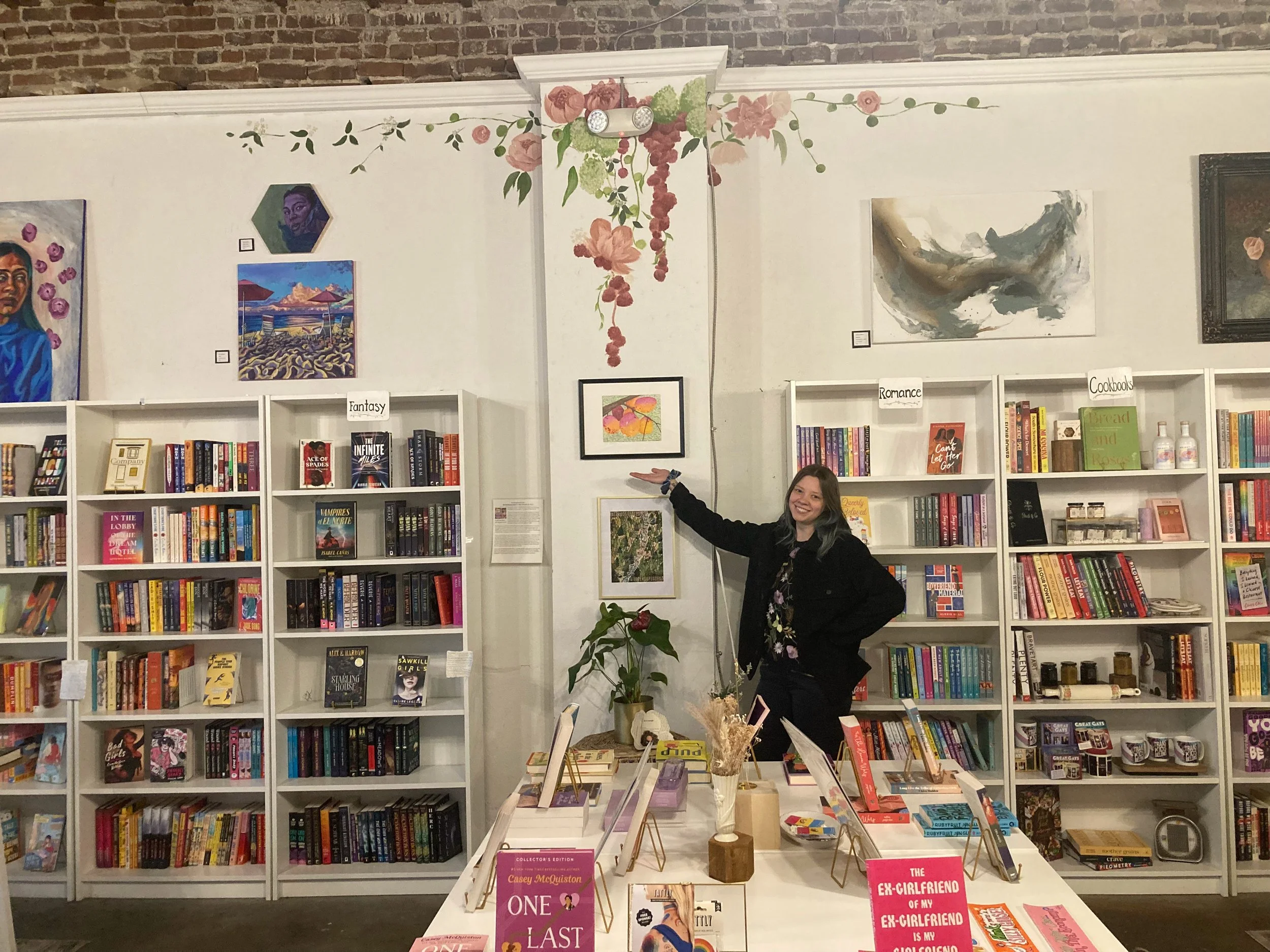 In a bookshop an artist is gesturing to a piece of artwork on the wall