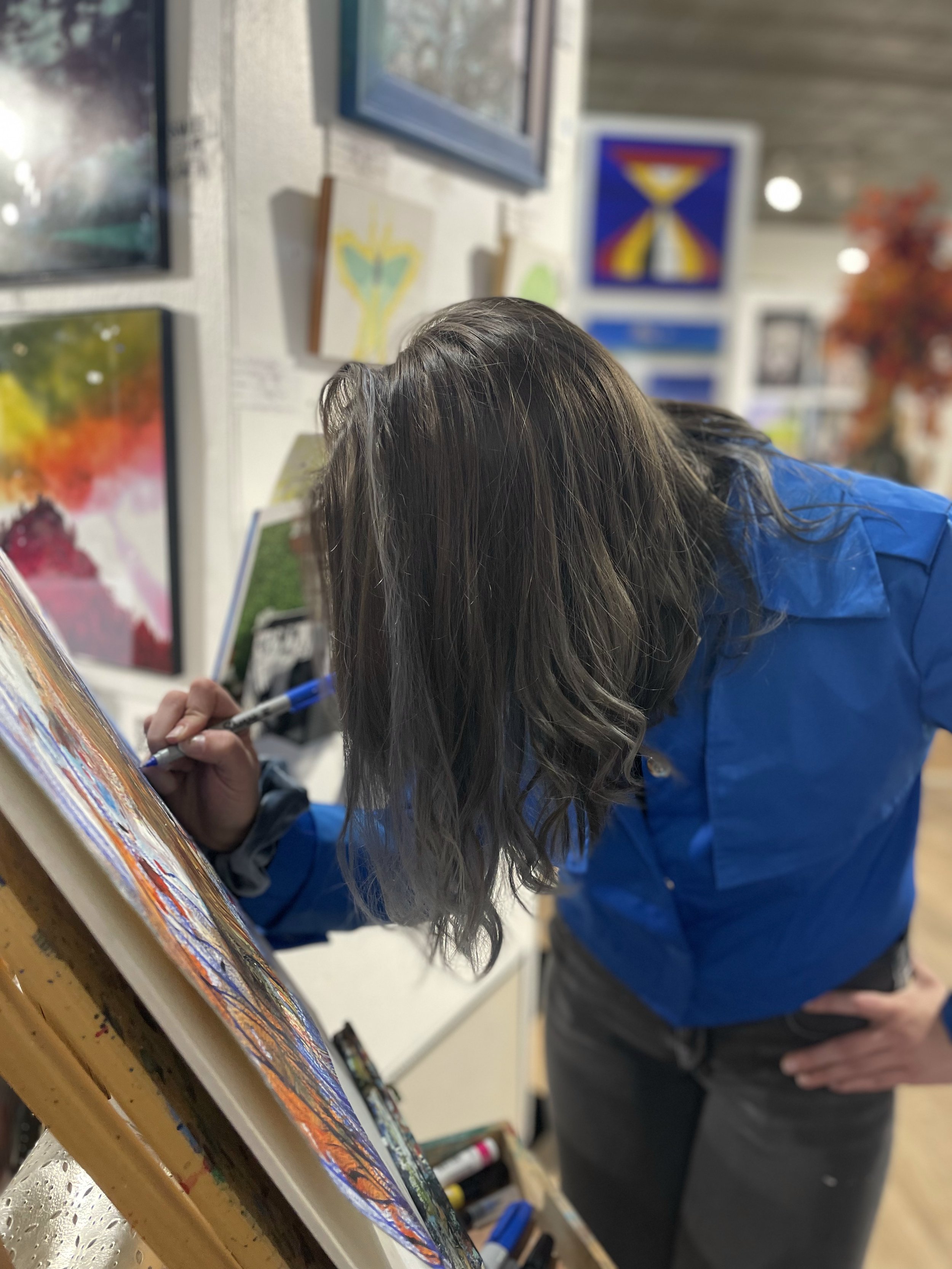 Artist with hair covering face wearing a blue jacket drawing at an easel. 