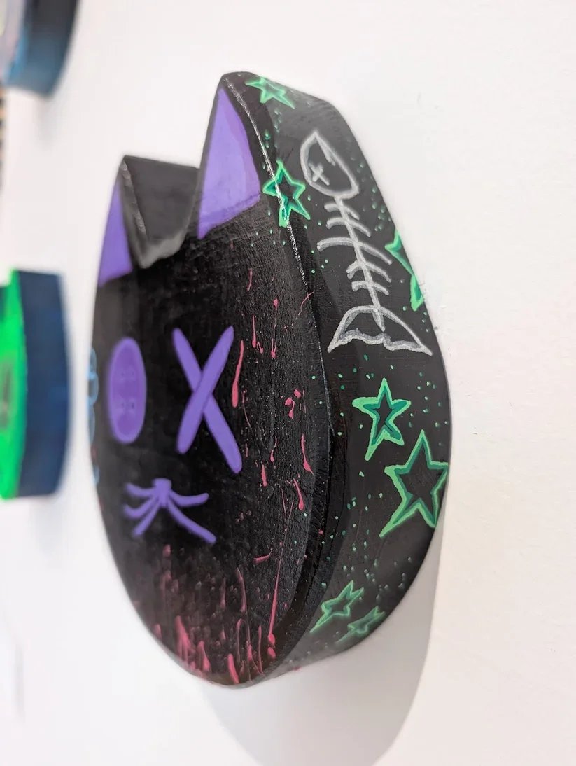 Painted wooden cat head art hanging on gallery wall. Cat head is black with purple eyes and mouth. With pink splatter, blue clouds, and fish bone details.