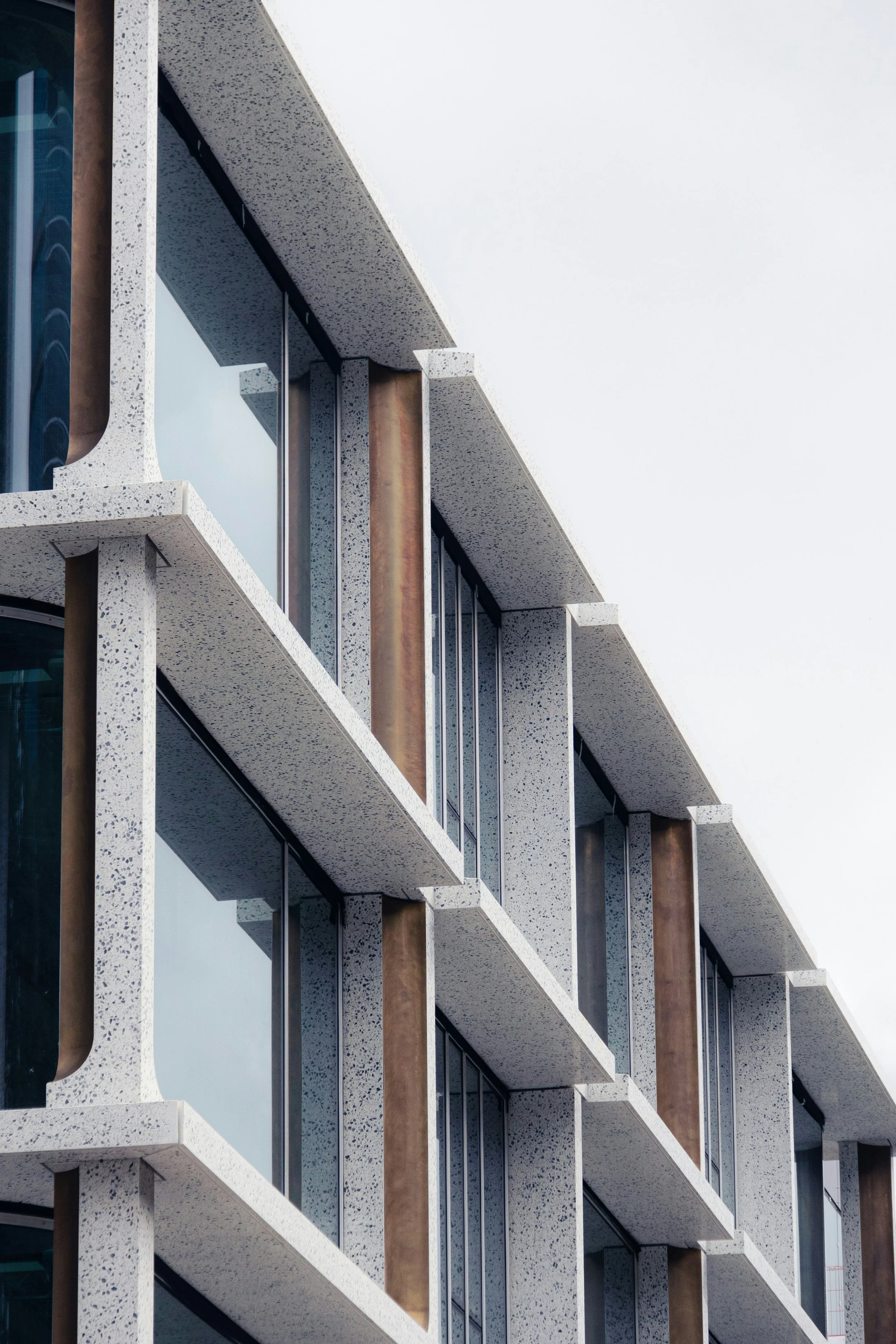 Close-up of a modern building's exterior with white speckled concrete balconies, glass windows, and vertical rust-colored metal panels, against a cloudy sky.