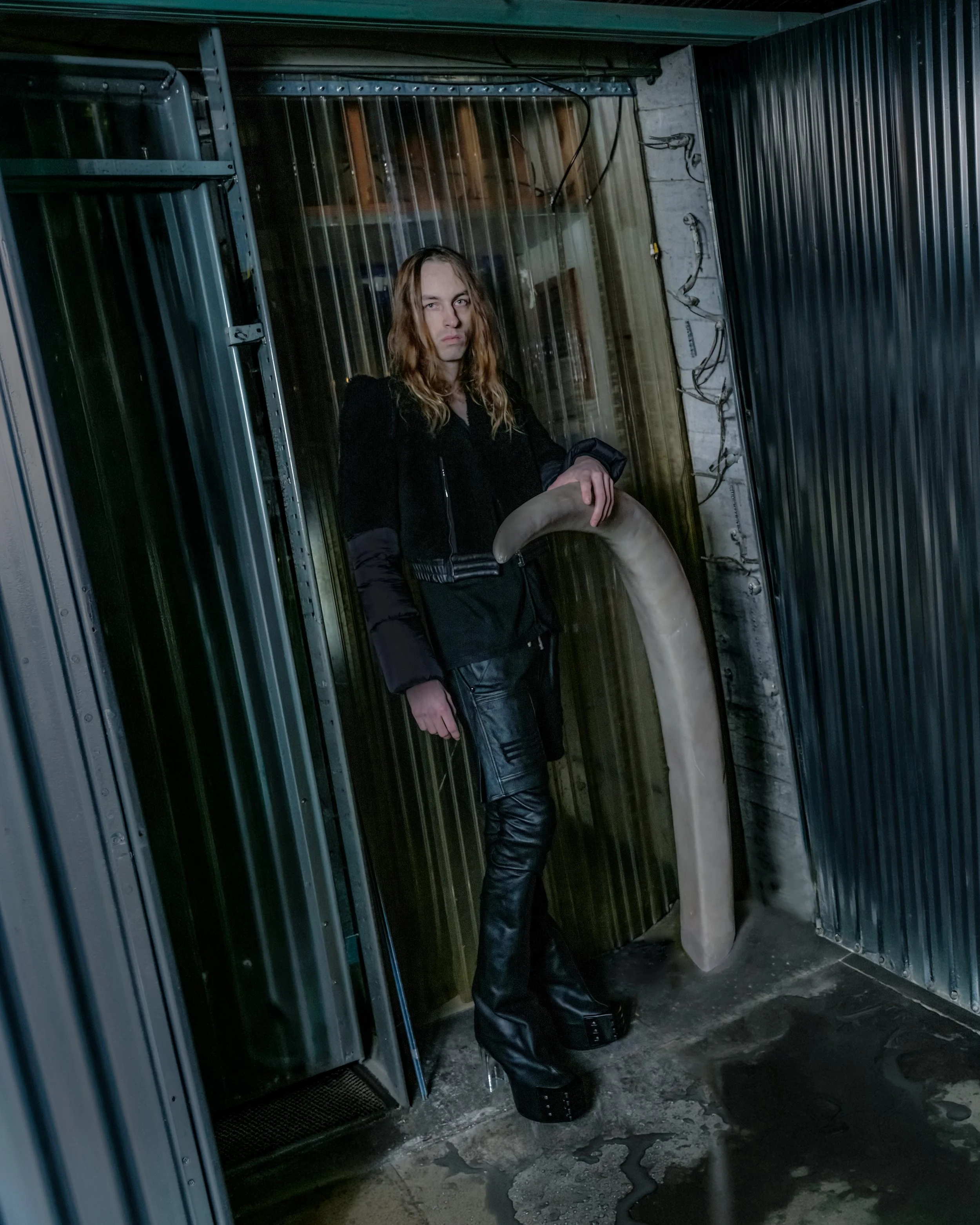 A person with long hair dressed in black leather pants and jacket standing inside a metallic elevator with reflective walls, holding a large, curved object resembling a horn.