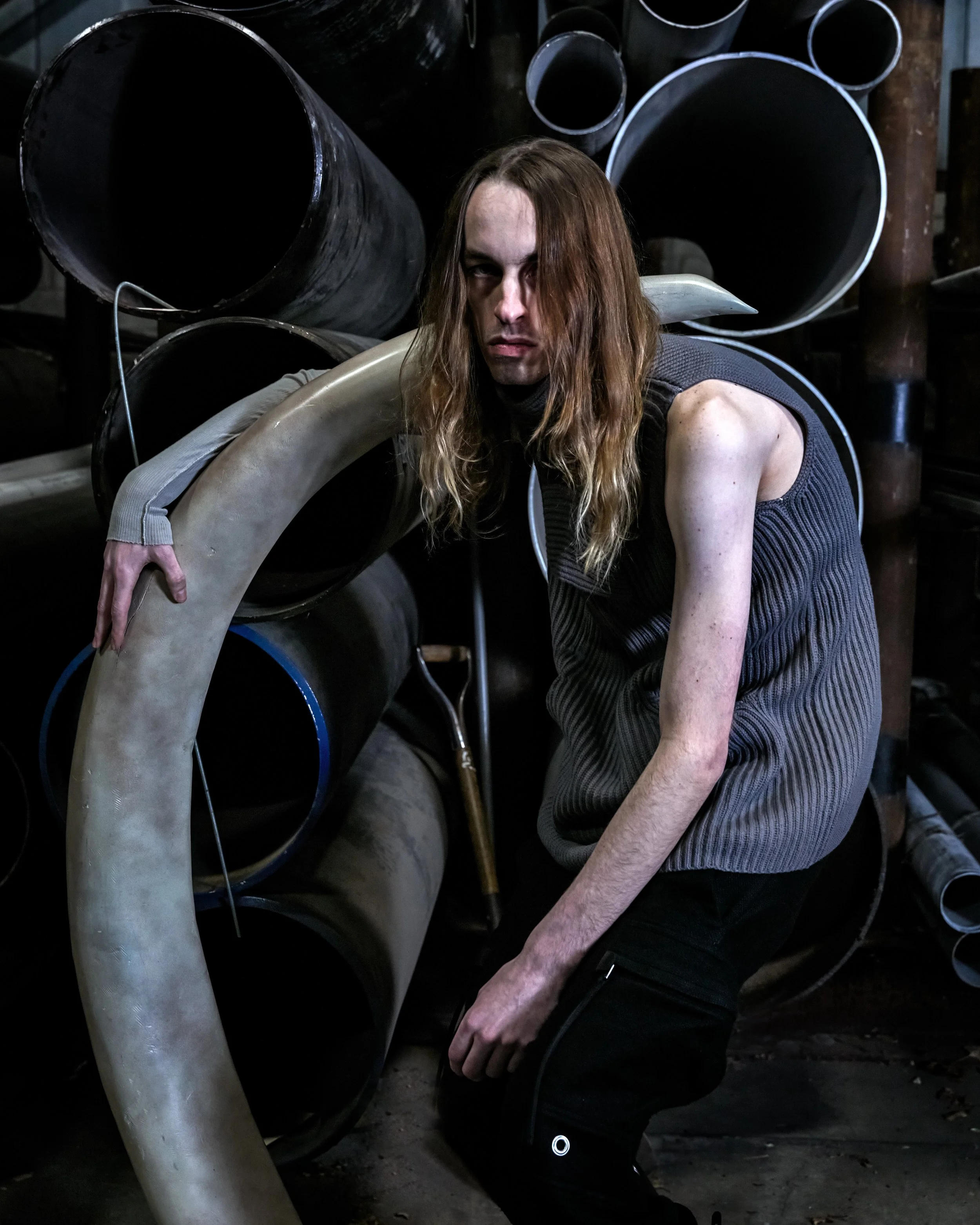 A person with long, wavy hair wearing a sleeveless gray shirt and black pants kneeling among large black and gray pipes in an industrial setting.
