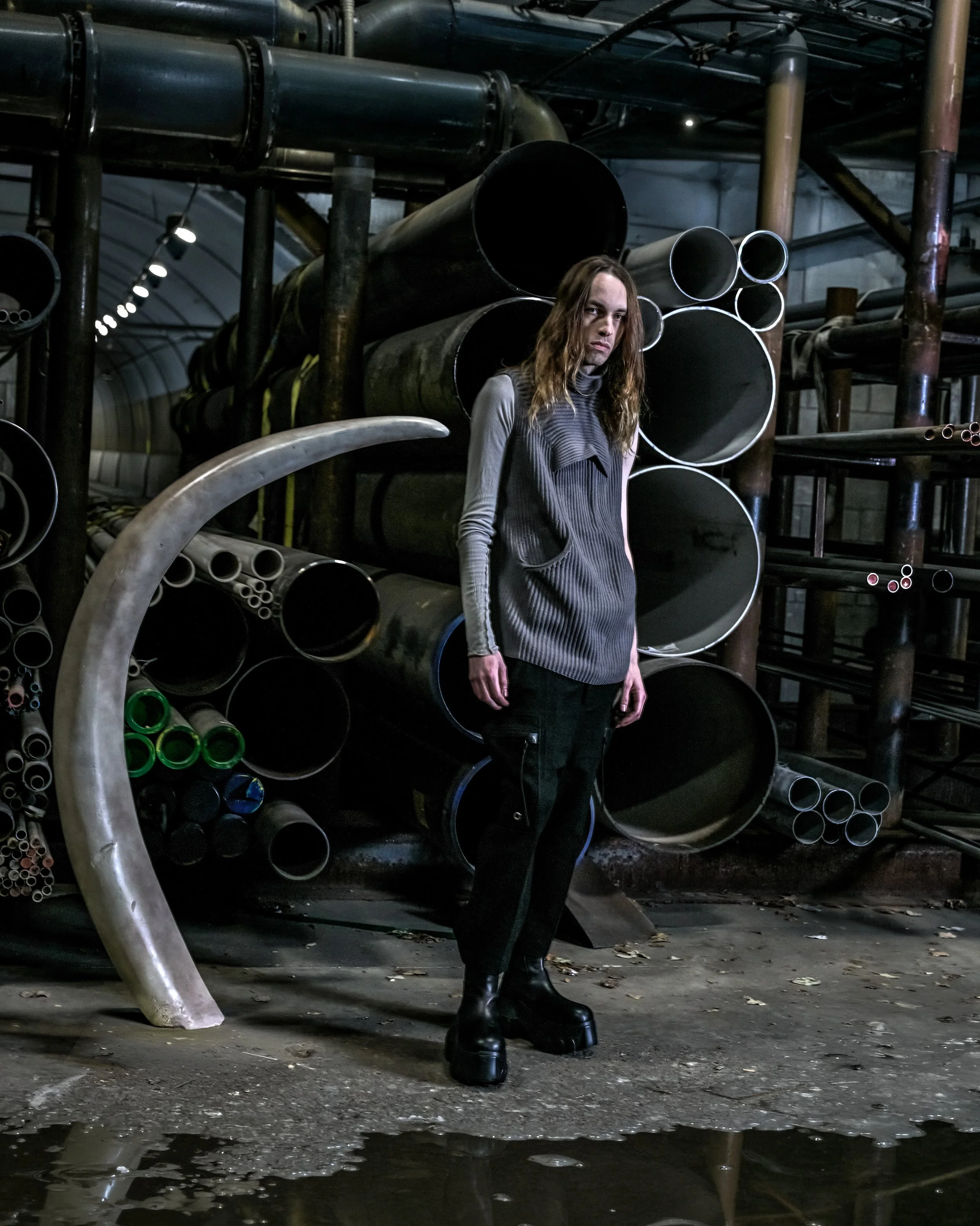 A woman with long hair wearing a gray and black outfit and platform shoes standing in an industrial setting with large pipes and metal structures.