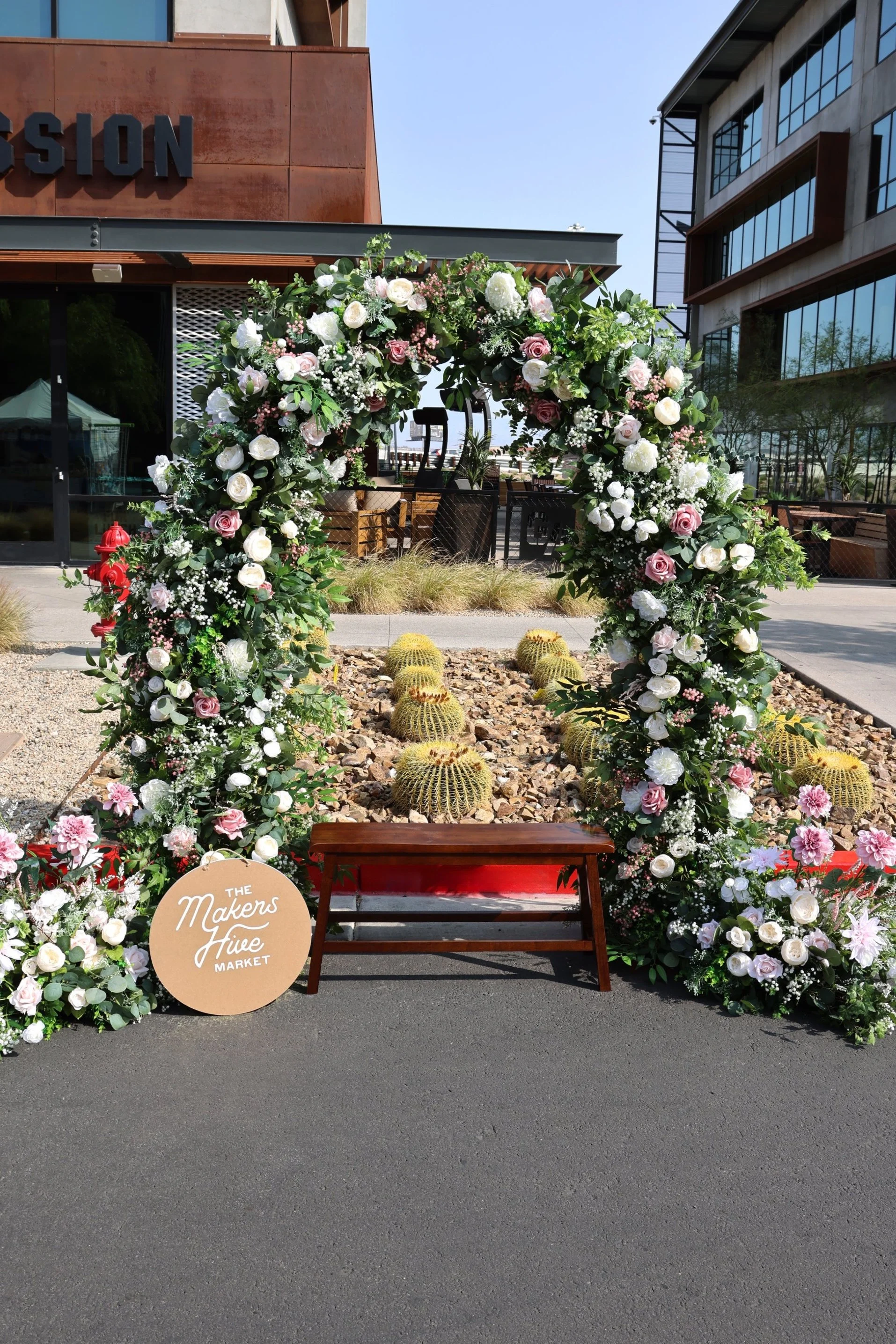 Pink and purple floral and greenery arch with a wooden bench in the center and a sign that reads "The Makers Hive Market"