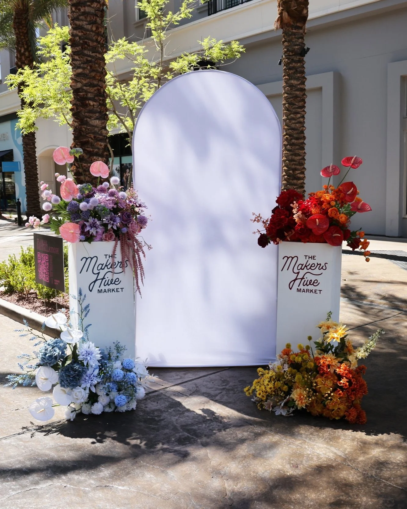 Four monochromatic faux floral arrangements with two white pedestal stands with a white arch fabric backdrop