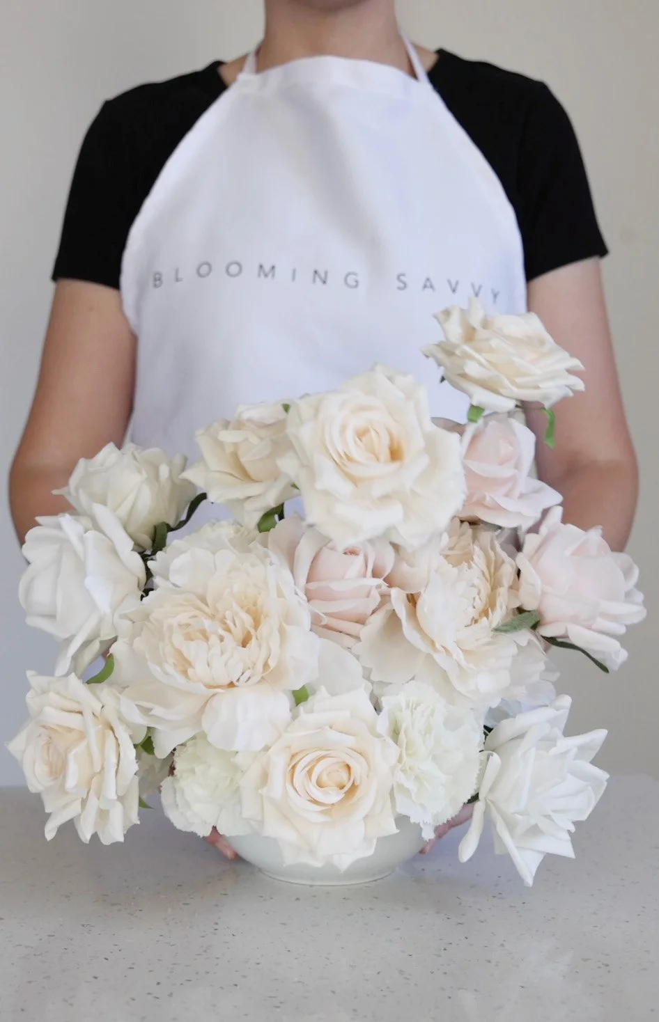 Person in white apron behind a white and cream rose floral arrangement in a white vase