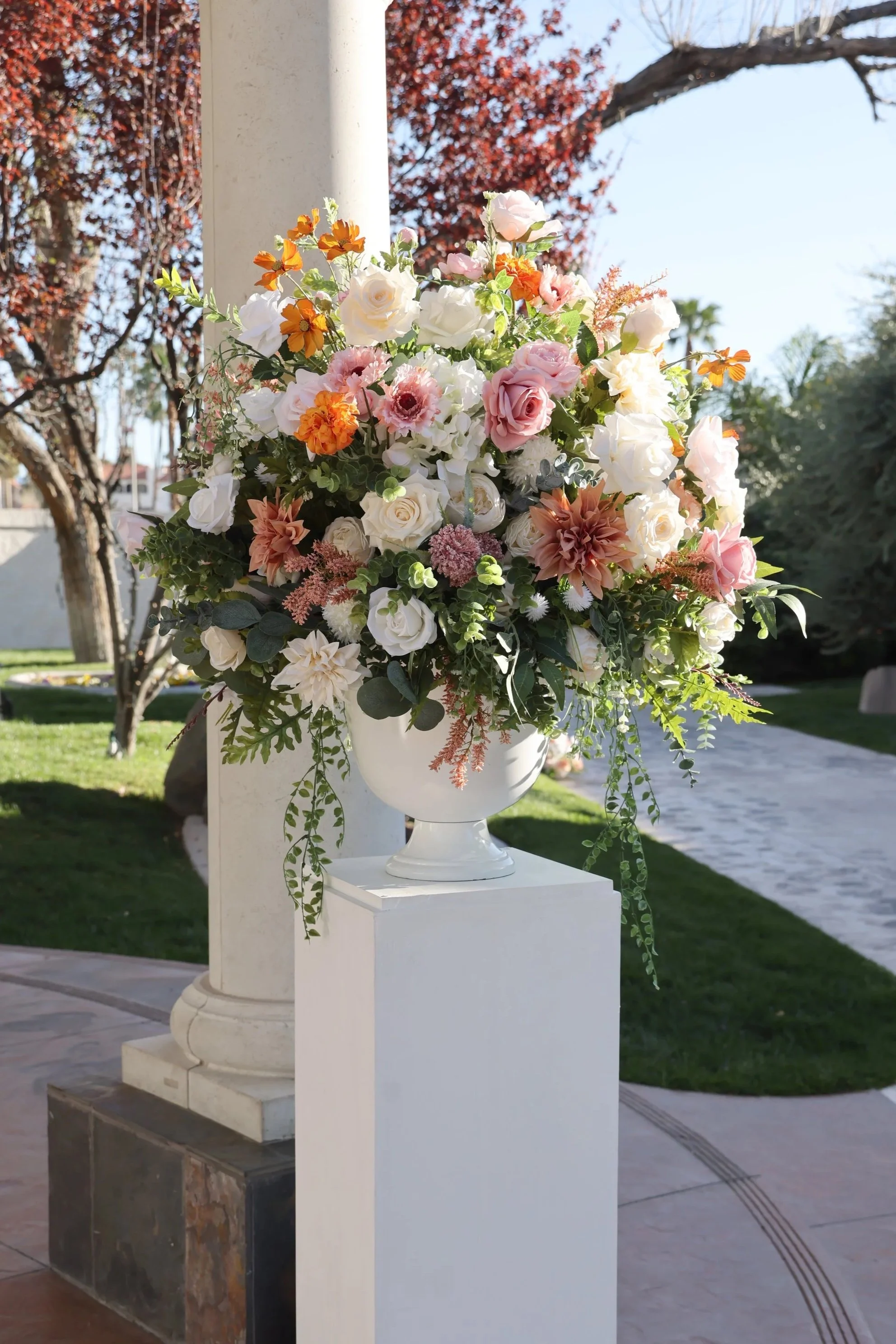 Urn vase arrangement with lush greenery and spring flowers on a white pedestal stand.