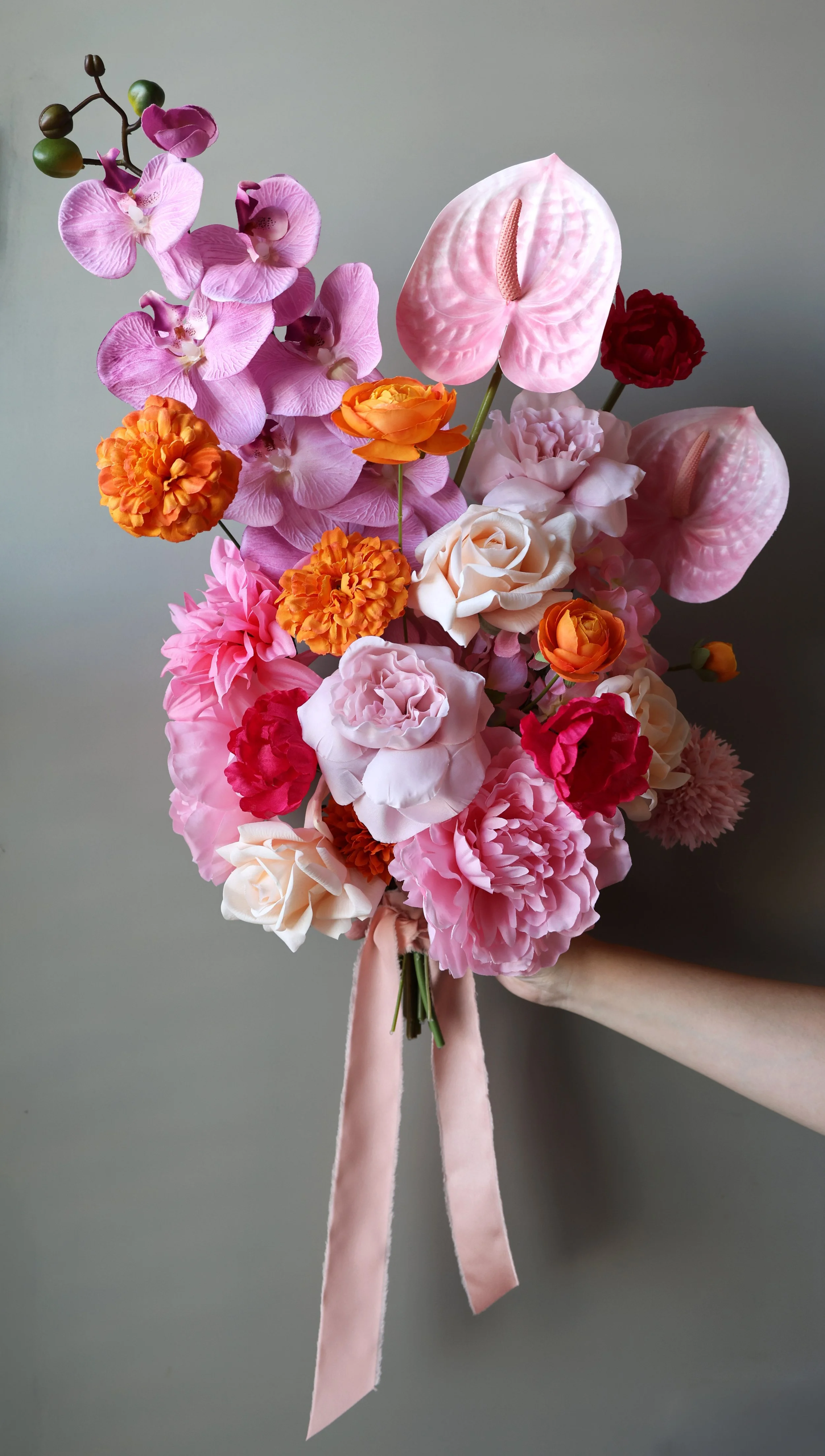 Hand holding a bright pink and orange bridal bouquet made with orchid, anthurium, peony, and rose flowers