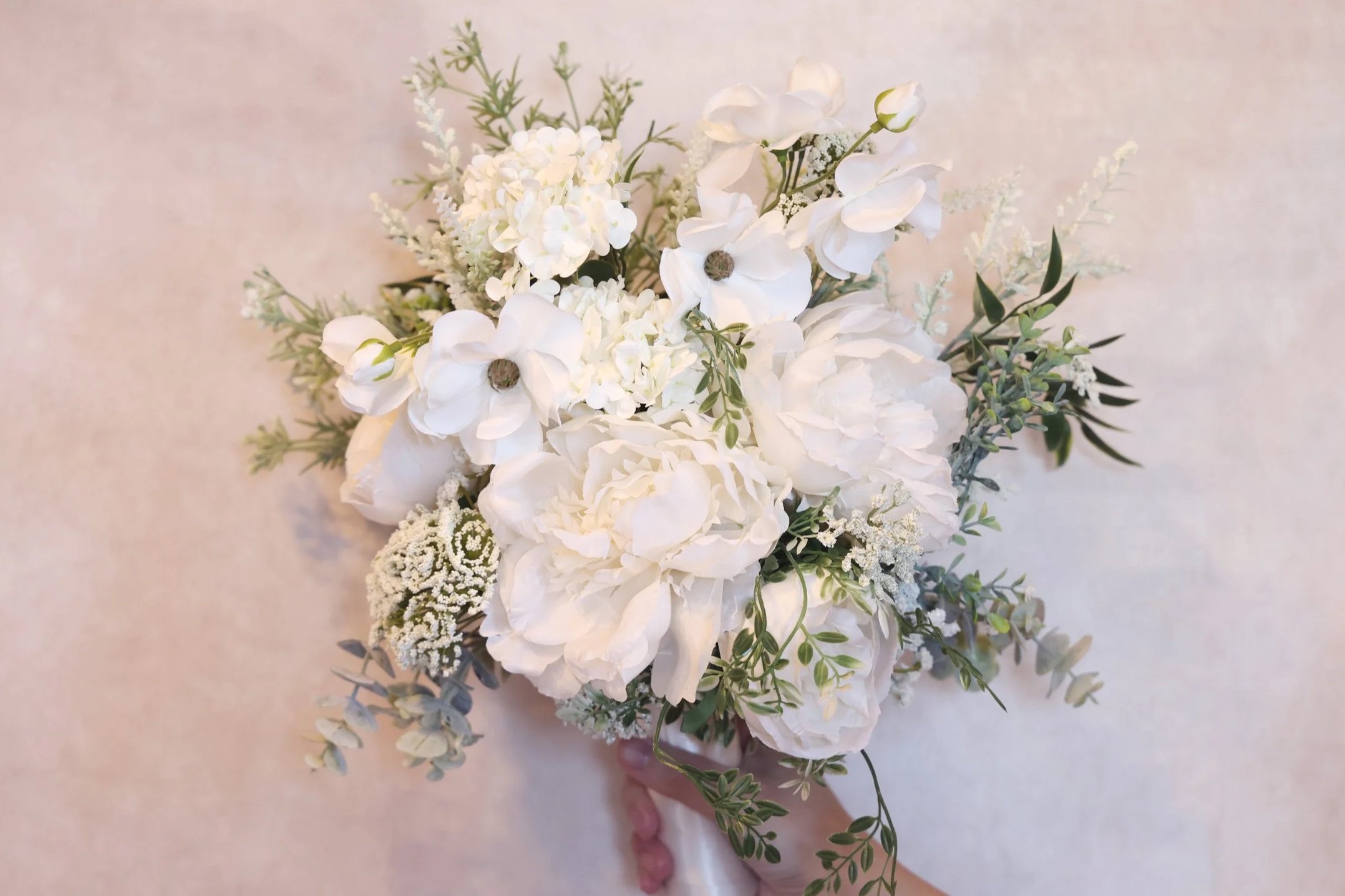 A white floral bouquet with peonies, hydrangeas, and greenery, held by a hand against a light-colored background.