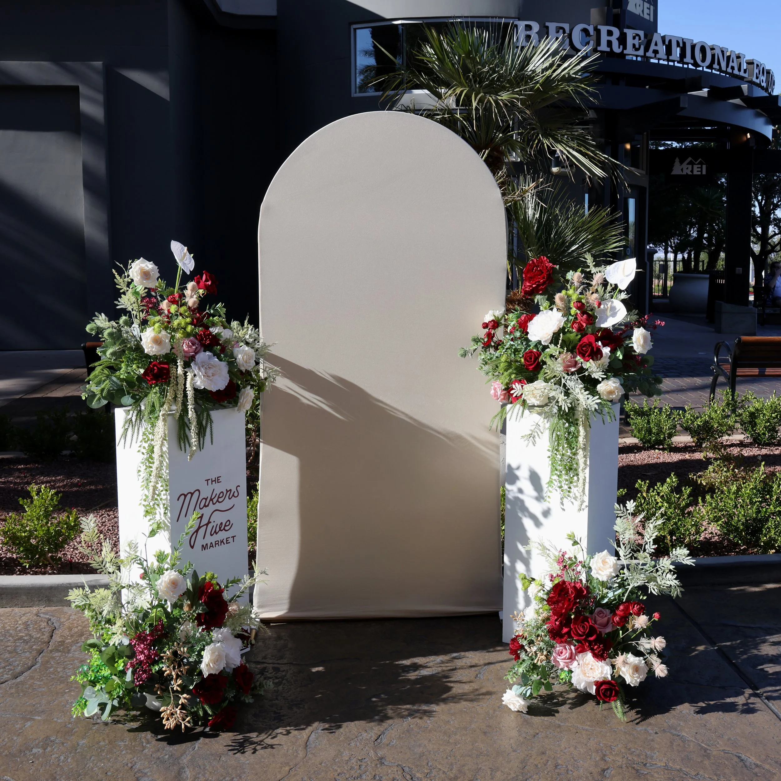 Flower photo op with four flower arrangements in burgundy, mauve, and beige colors mixed with greenery with two white pedestal stands and a beige fabric arch backdrop