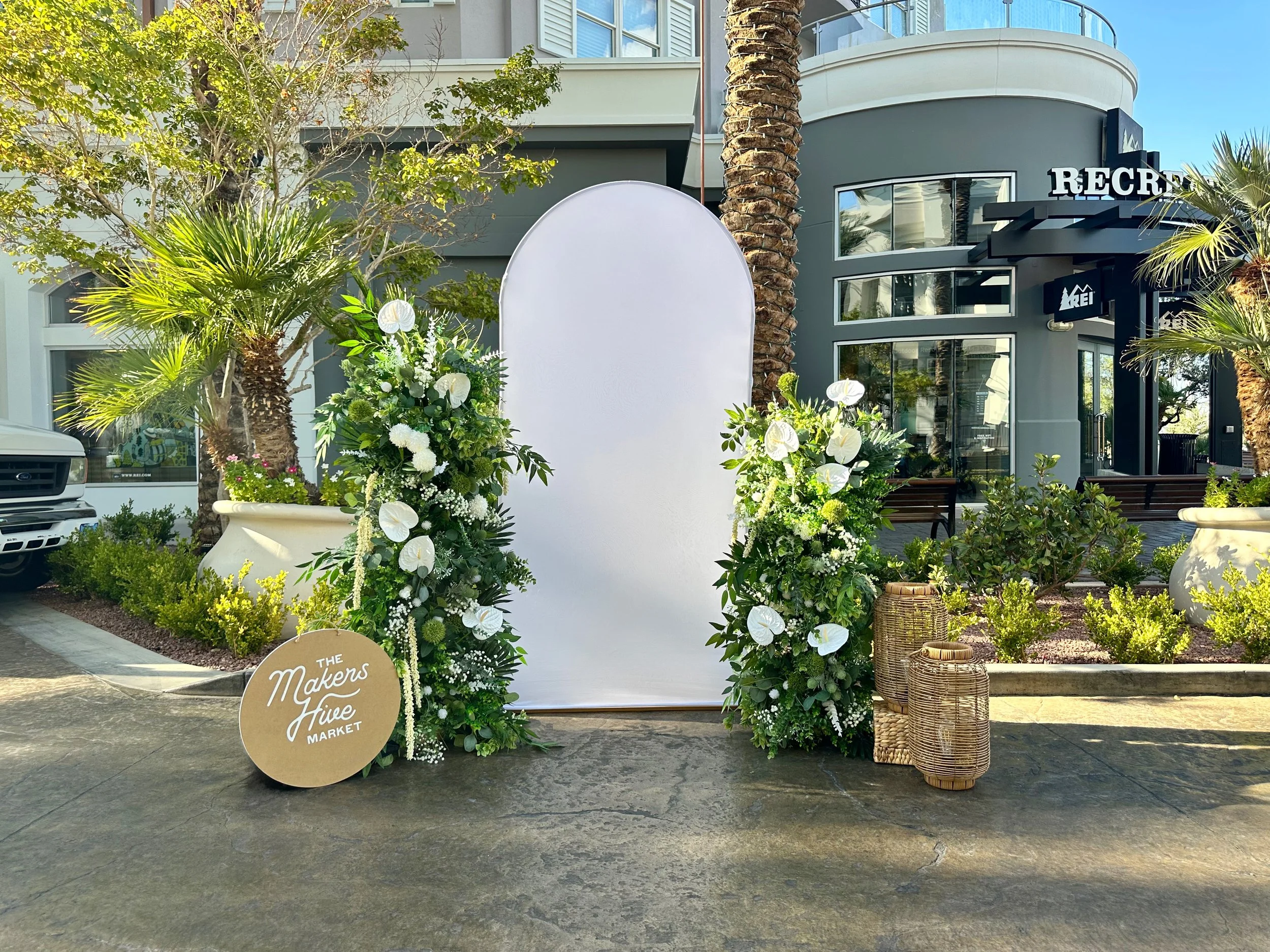 Tropical flower and greenery photo op with greenery pillars with white anthurium blooms and amaranthus with a white fabric arch backdrop and beige lanterns and a sign that reads "The Makers Hive Market"