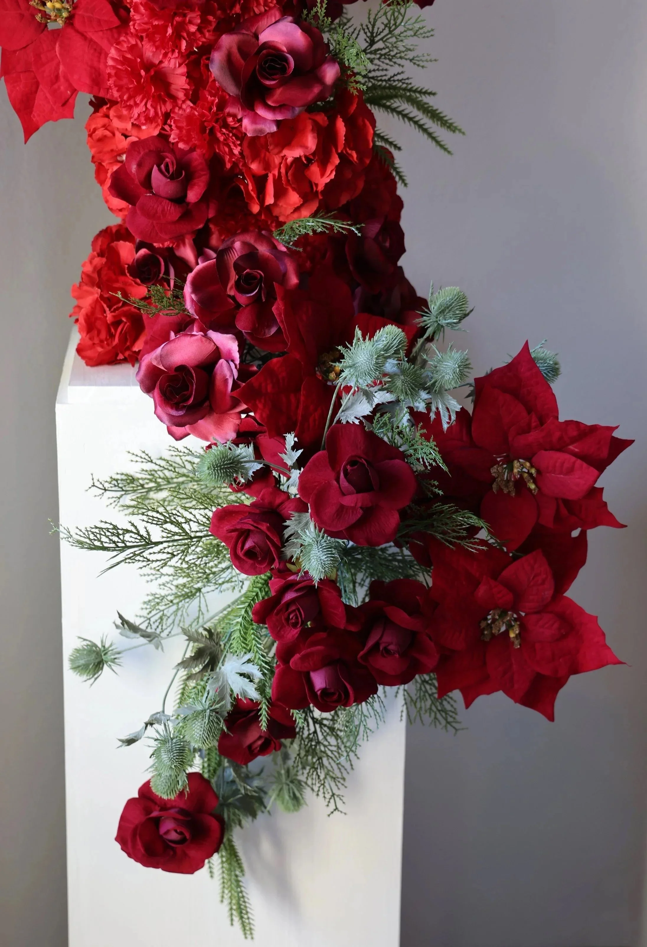 Christmas themed flower arrangement sculpture on a white pedestal stands made with red poinsettia, roses, and hydrangea florals and pine needle greenery accents
