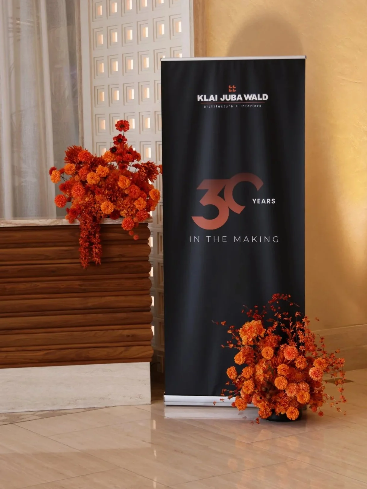 Two large monochromatic orange floral arrangements are shown, one on top of a beige wooden counter and the other at the base of a black tall banner that reads "Klai Juda Wald Architecture + Interirors 30 Years in the Making"
