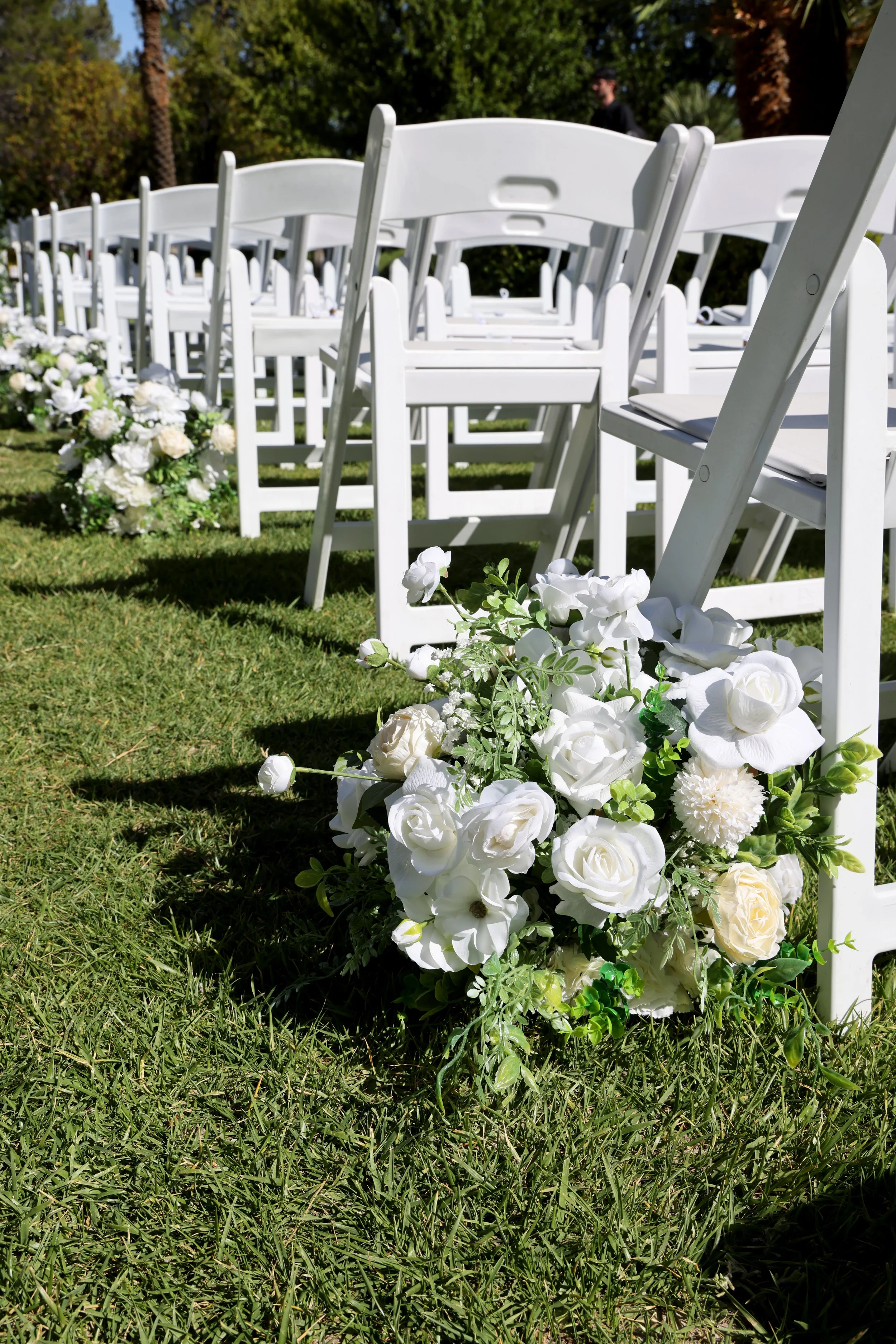 Greenery and white floral arrangements on grass next to white chairs lining wedding ceremony aisle