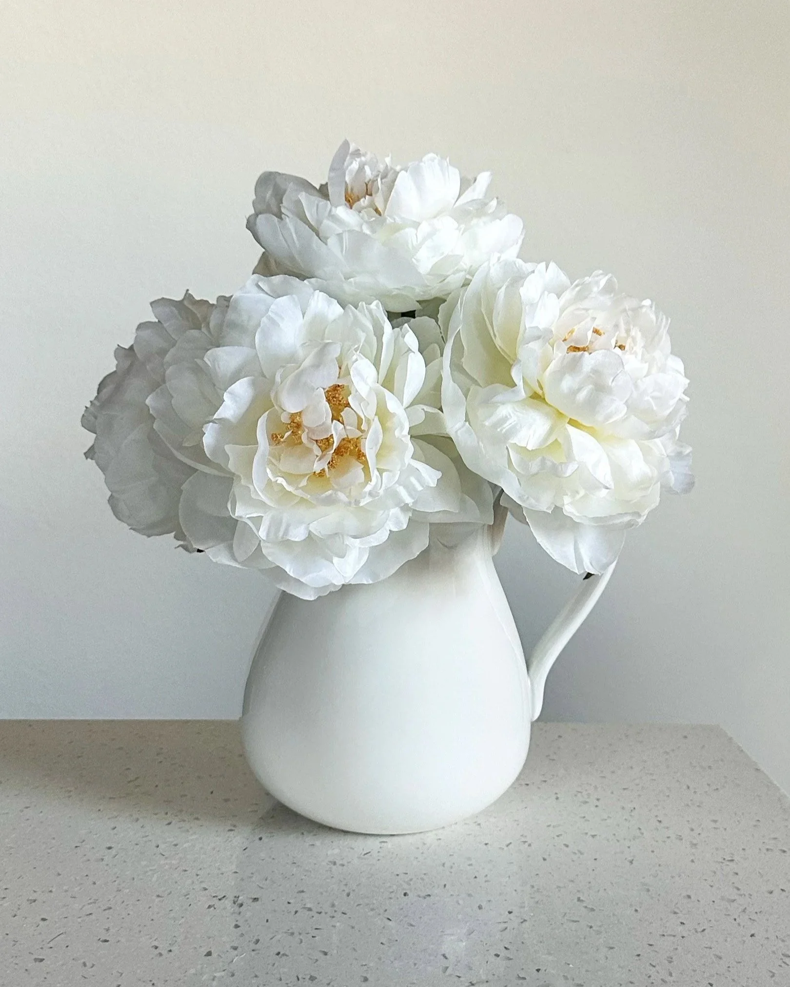 Artificial ivory peonies in a white ceramic pitcher on a white stone counter