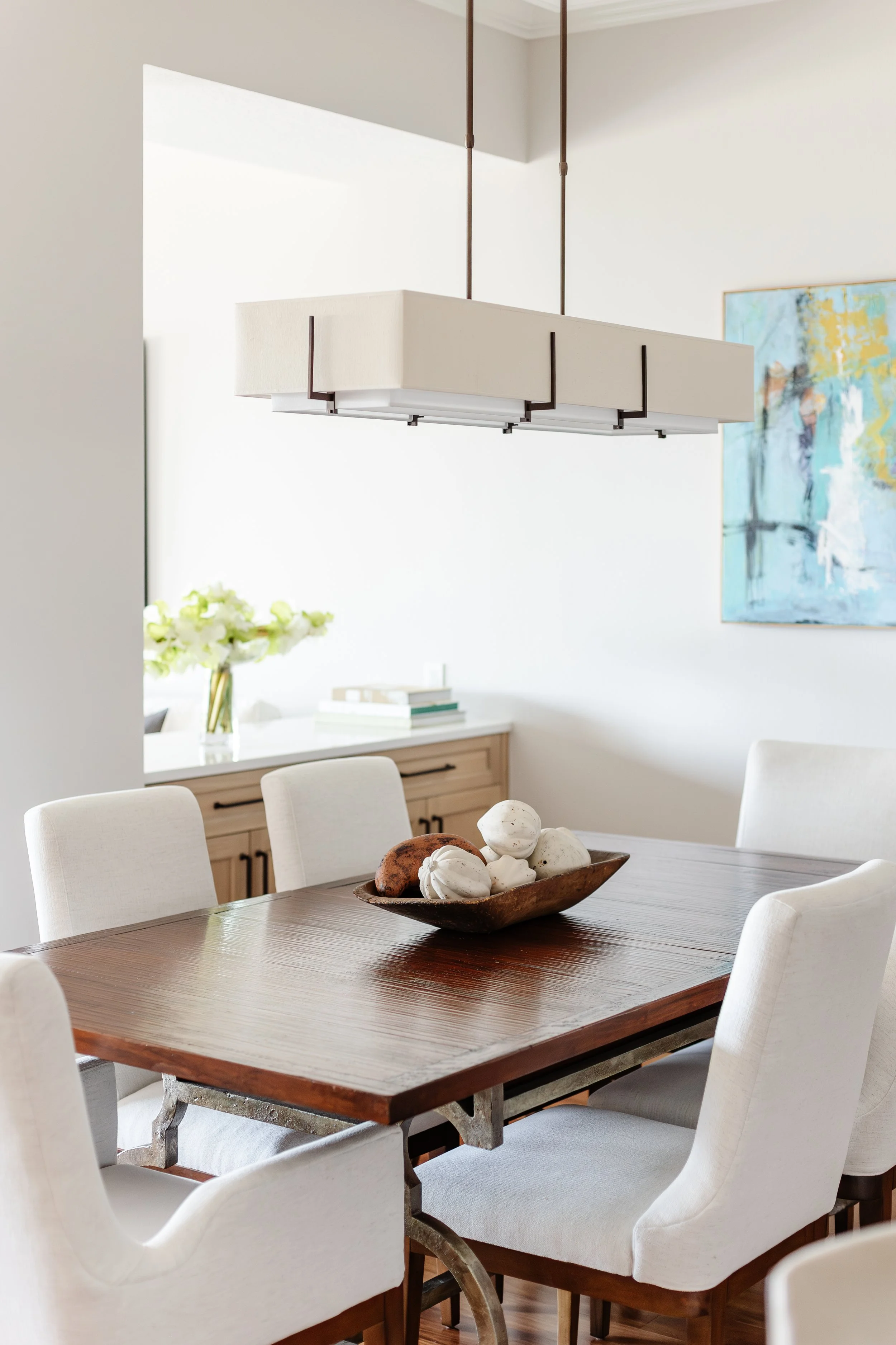 A modern dining room with a wooden table, white upholstered chairs, a contemporary pendant light, a sideboard with books and flowers, and an abstract painting on the wall.