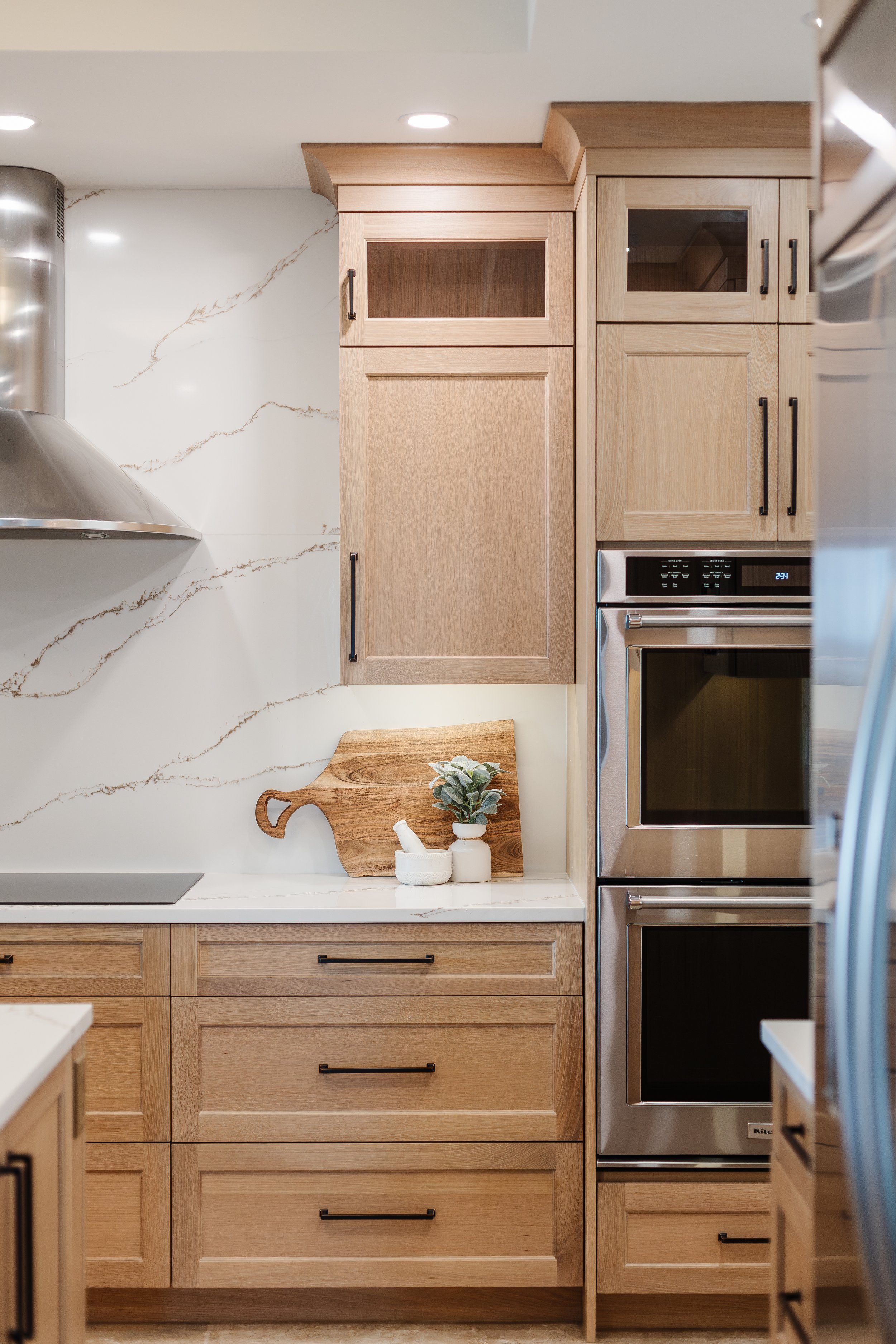 Modern kitchen with light wood cabinets, a marble countertop, and stainless steel double oven. Decor includes a wooden cutting board, a small plant, and a mortar and pestle.