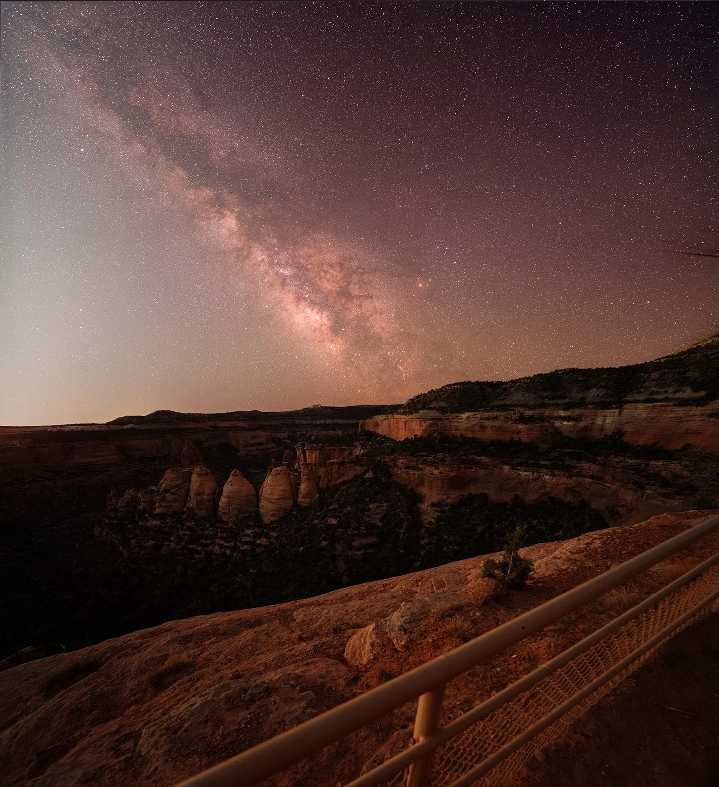 A nighttime view of the Milky Way galaxy over a canyon, with a viewing platform and railing in the foreground.