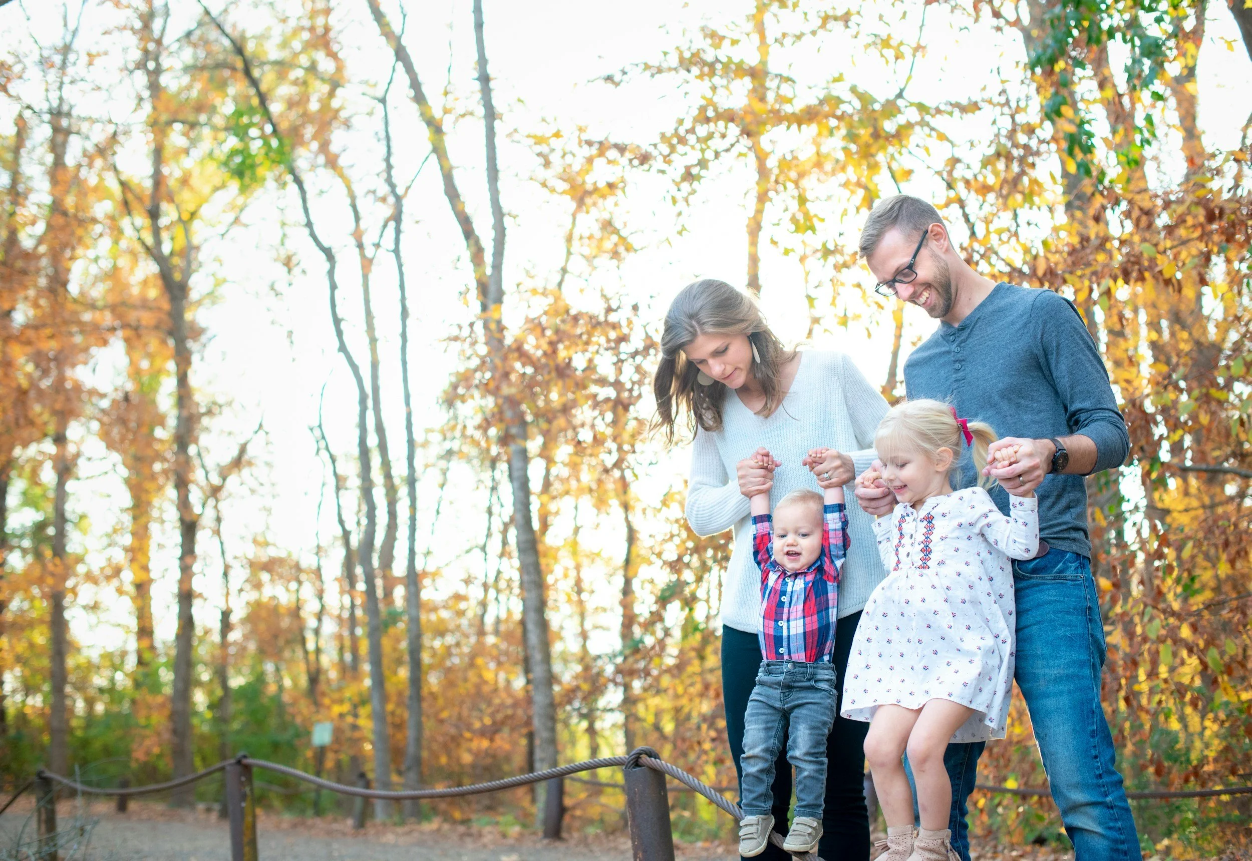 Family of four holding hands and smiling in a forest during autumn.
