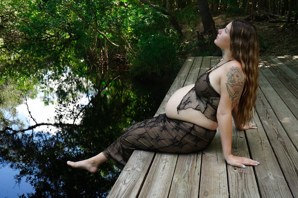 Pregnant woman sitting on a wooden deck by a pond, wearing a sheer dress, with trees and water in the background.