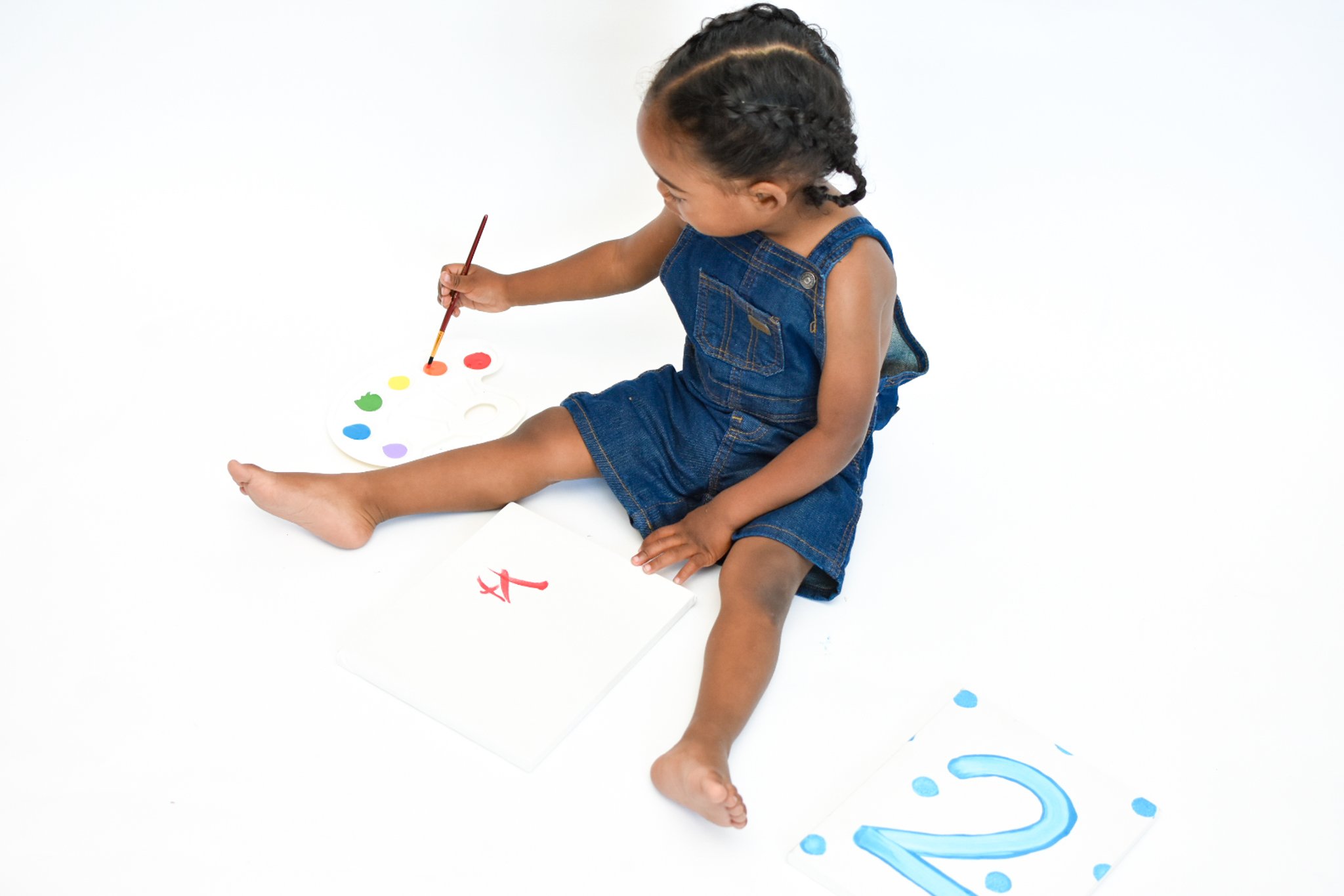 Child in denim overalls painting with a brush on a canvas, colorful palette next to them.