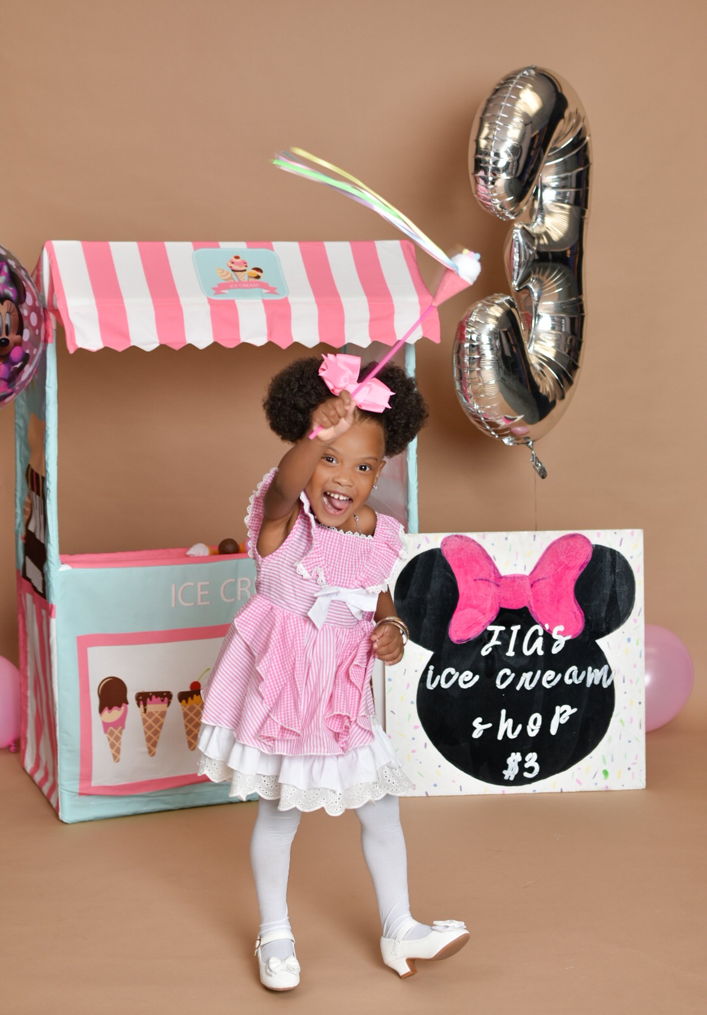 Young girl in a pink dress with pink bow, holding a wand, stands near a play ice cream shop with striped awning and Minnie Mouse-themed sign. Silver number three balloon in background.