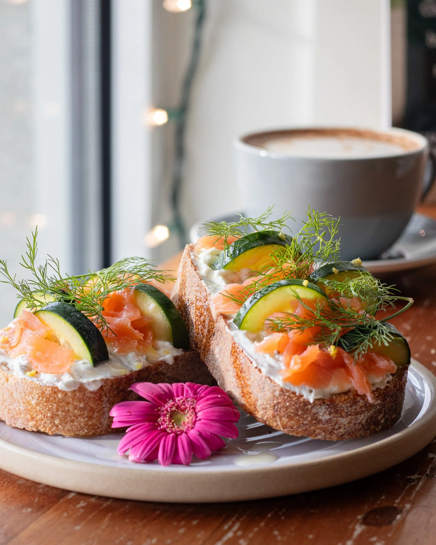 Open-faced sandwich from BellDen Cafe with salmon, cucumber, dill, and cream cheese on toasted bread, garnished with a pink flower and served with a cup of coffee in the background.