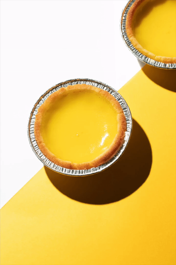 Two egg custard tarts in aluminum foil cups on a surface split diagonally between white and yellow, with shadows cast behind them.