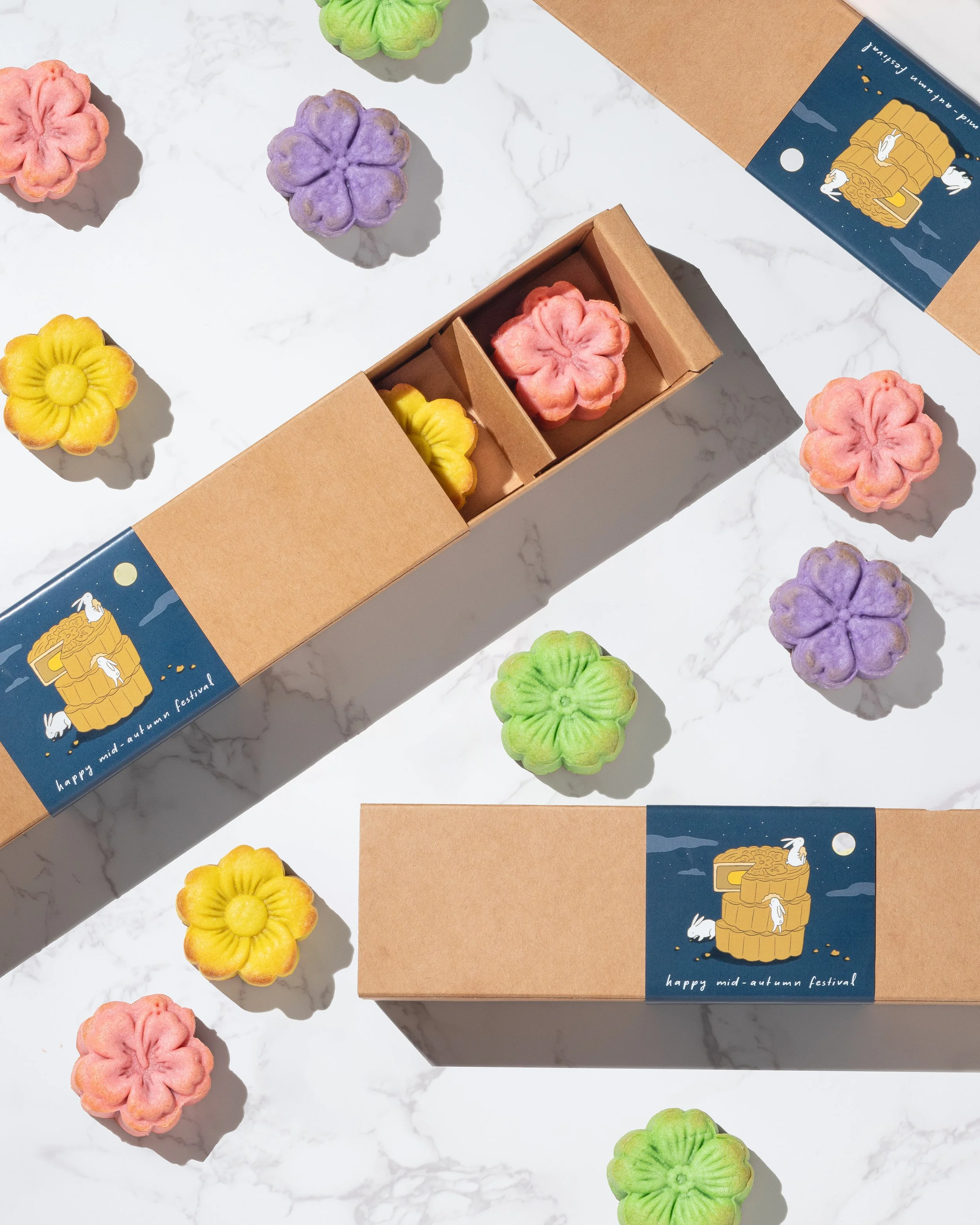 Colorful flower-shaped mooncakes from Kemi Dessert Bar on a white marble surface. There are alsomooncakes in a brown cardboard box with a blue label that says 'happy mid-autumn festival' and features illustrations of a moon and rabbits.