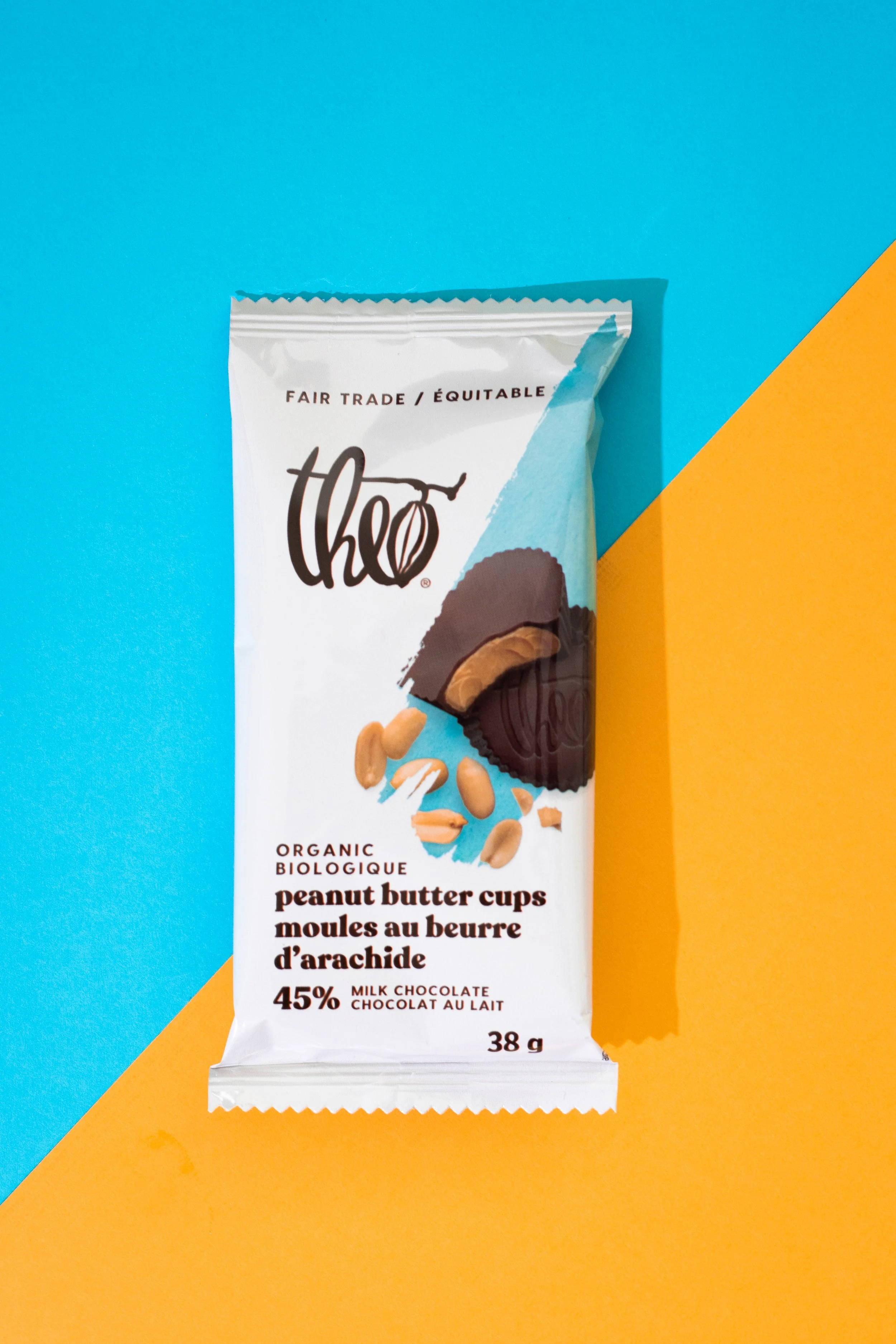 A chocolate bar from Theo Chocolate in a white wrapper labeled 'Peanut Butter Cups' with text indicating it is organic, fair trade, milk chocolate with 45% cocoa, weighing 38 grams, placed against a backdrop divided diagonally into bright blue and ye