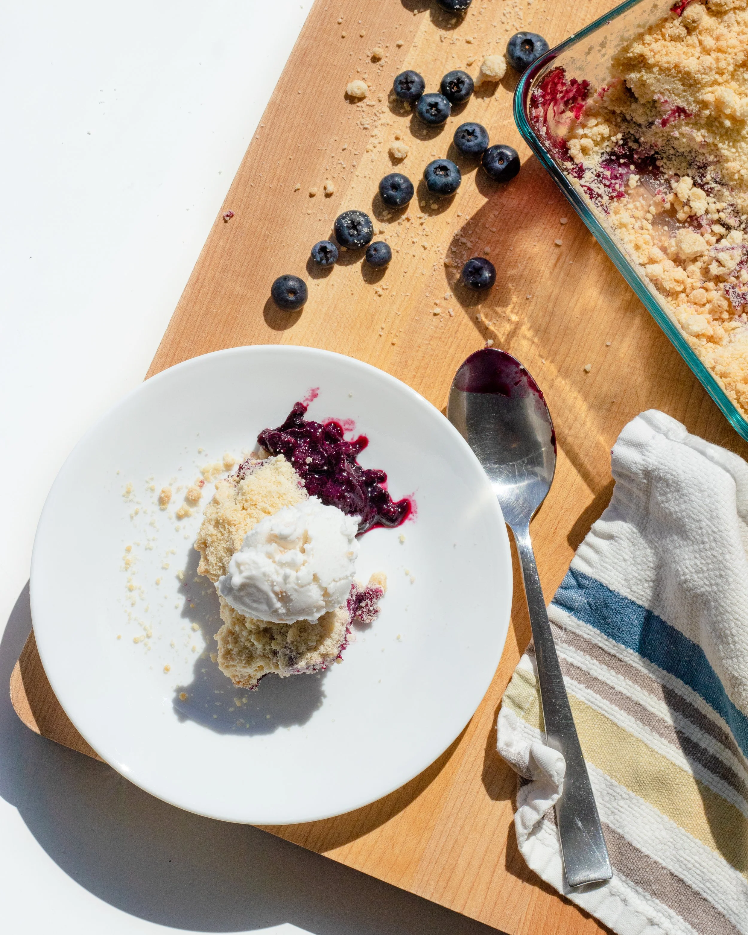 A partially eaten serving of blueberry crumble with a scoop of vanilla ice cream on a white plate, placed on a wooden cutting board with scattered blueberries and crumble topping, next to a knife and a striped kitchen towel.