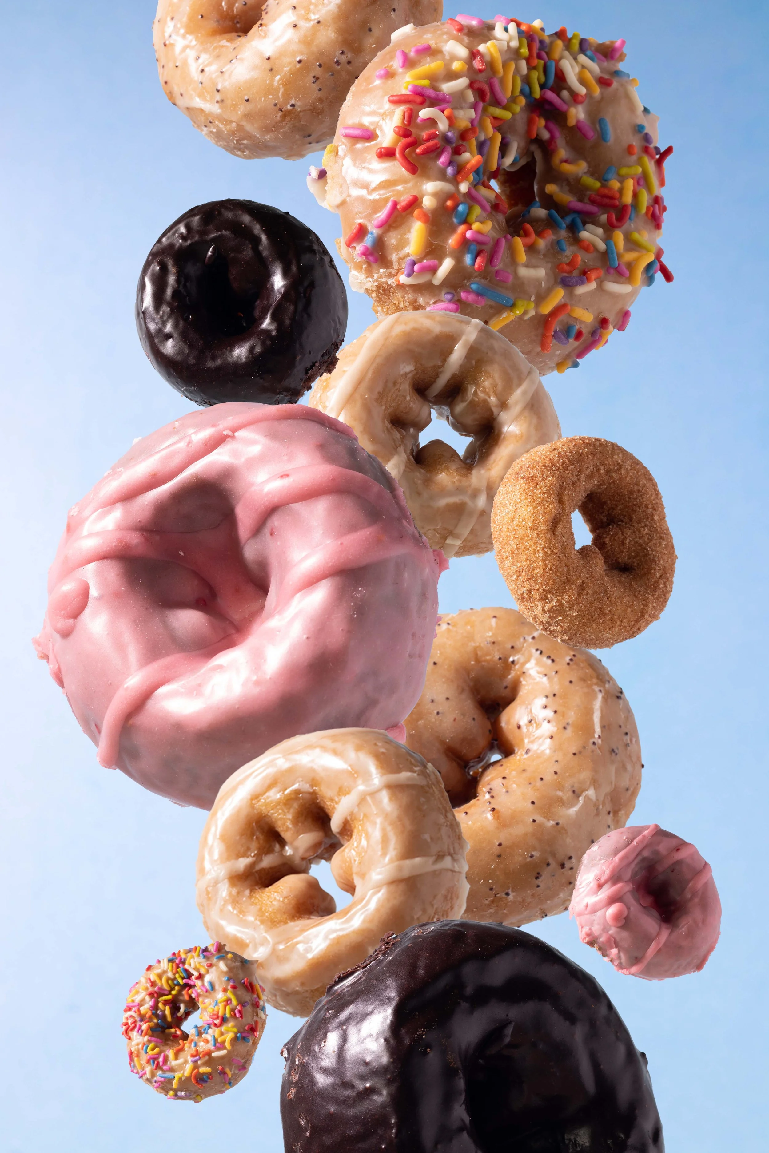 A vertical arrangement of assorted donuts from Might-O with different toppings, including chocolate, pink glaze, sprinkles, and caramel, against a light blue background.