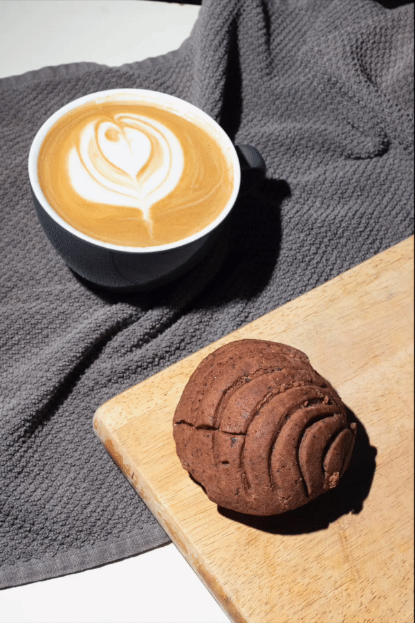 A cup of coffee with latte art next to a chocolate concha bread on a wooden board, placed on a gray textured cloth.