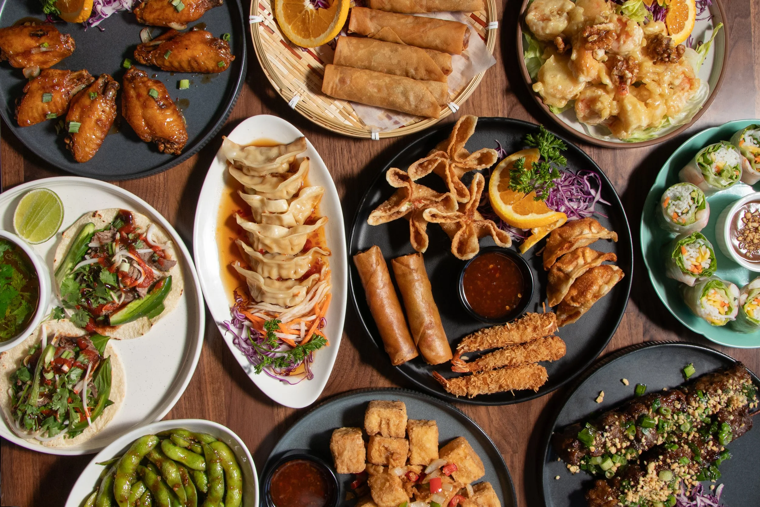 Flatlay of Asian dishes from Lotus on the Beach in West Seattle, including spring rolls, dumplings, fried chicken wings, vegetable rolls, edamame, salad, and tofu served on various plates and bowls.