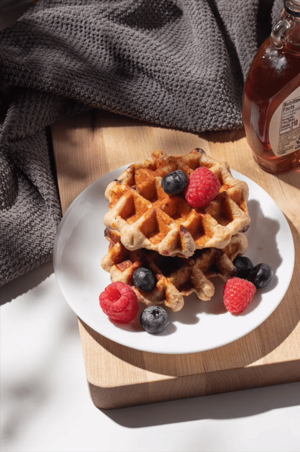 Two Belgian waffles topped with mixed berries on a white plate, placed on a wooden board.