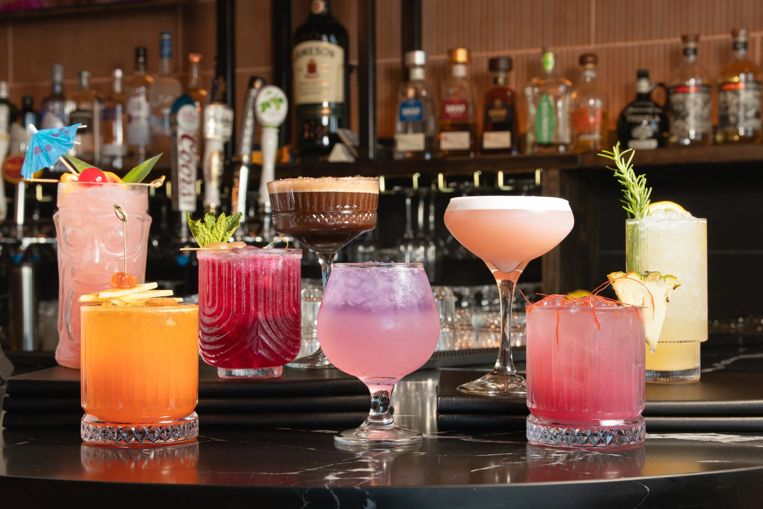 A variety of colorful cocktails from Lotus on the Beach in West Seattle on a bar counter, with a wooden bar backdrop featuring various liquor bottles.