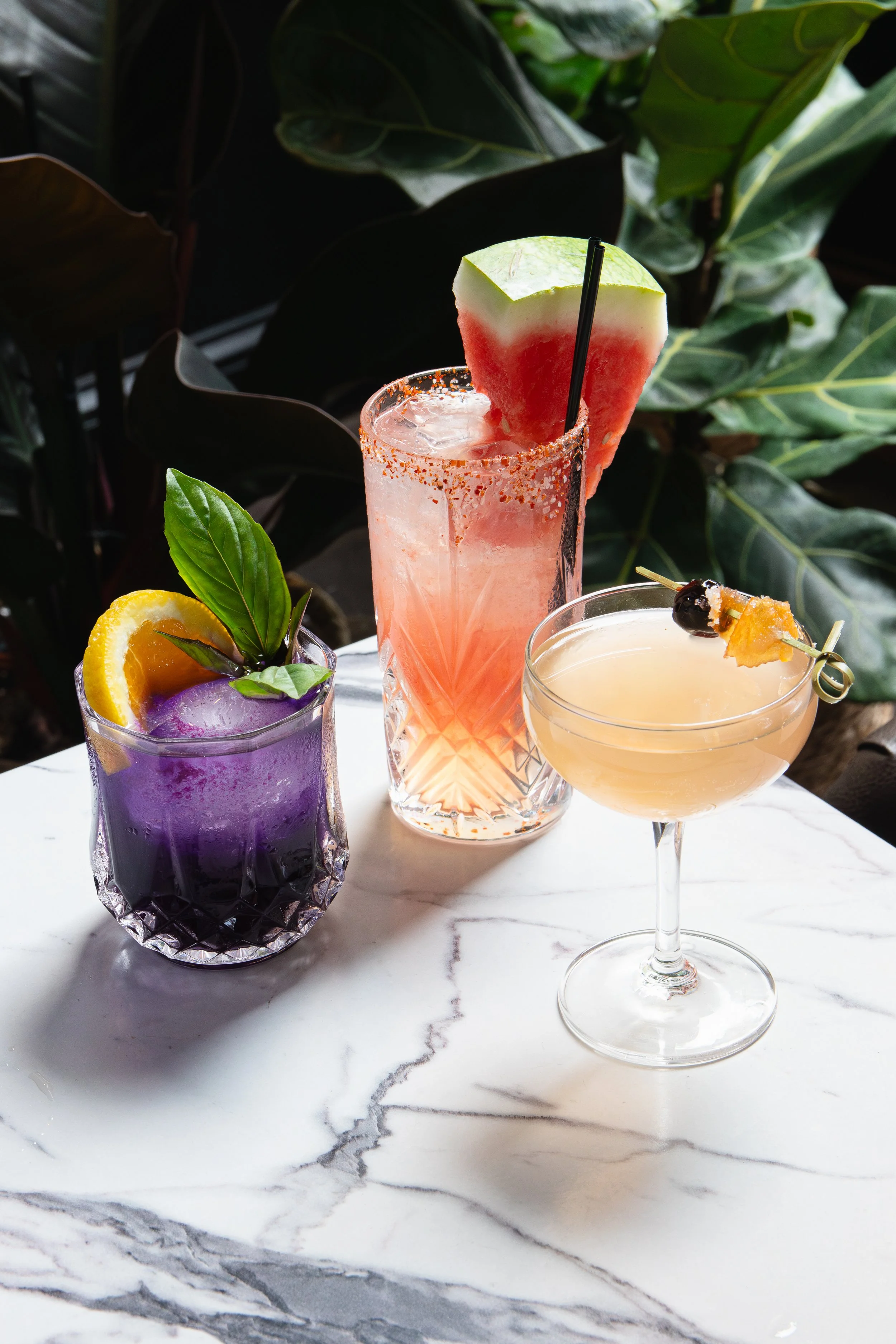 Three colorful cocktails from Kalabaw Seattle on a white marble table with green foliage in the background. One purple drink garnished with orange and basil, a pink watermelon-flavored drink with a watermelon slice, and a yellowish cocktail with a ch